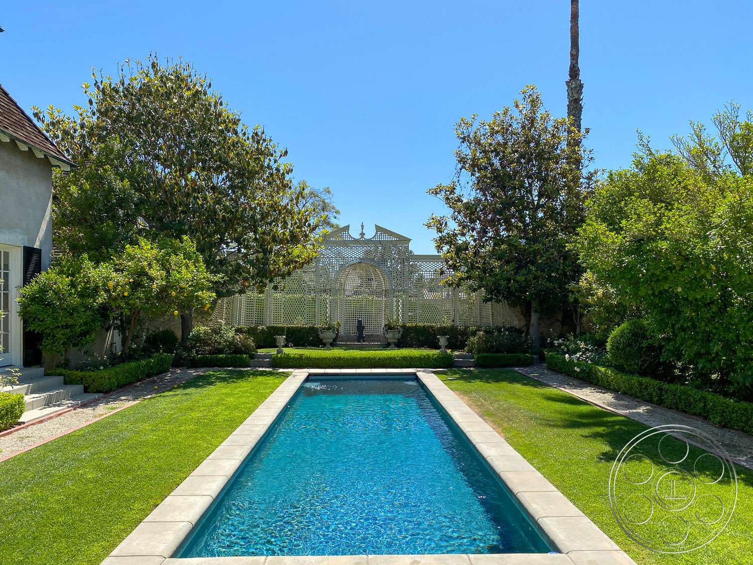 Long rectangular pool between clipped hedges and lawn, closed at the far end by a white lattice garden pavilion, at The French Chateau, a Hancock Park filming location