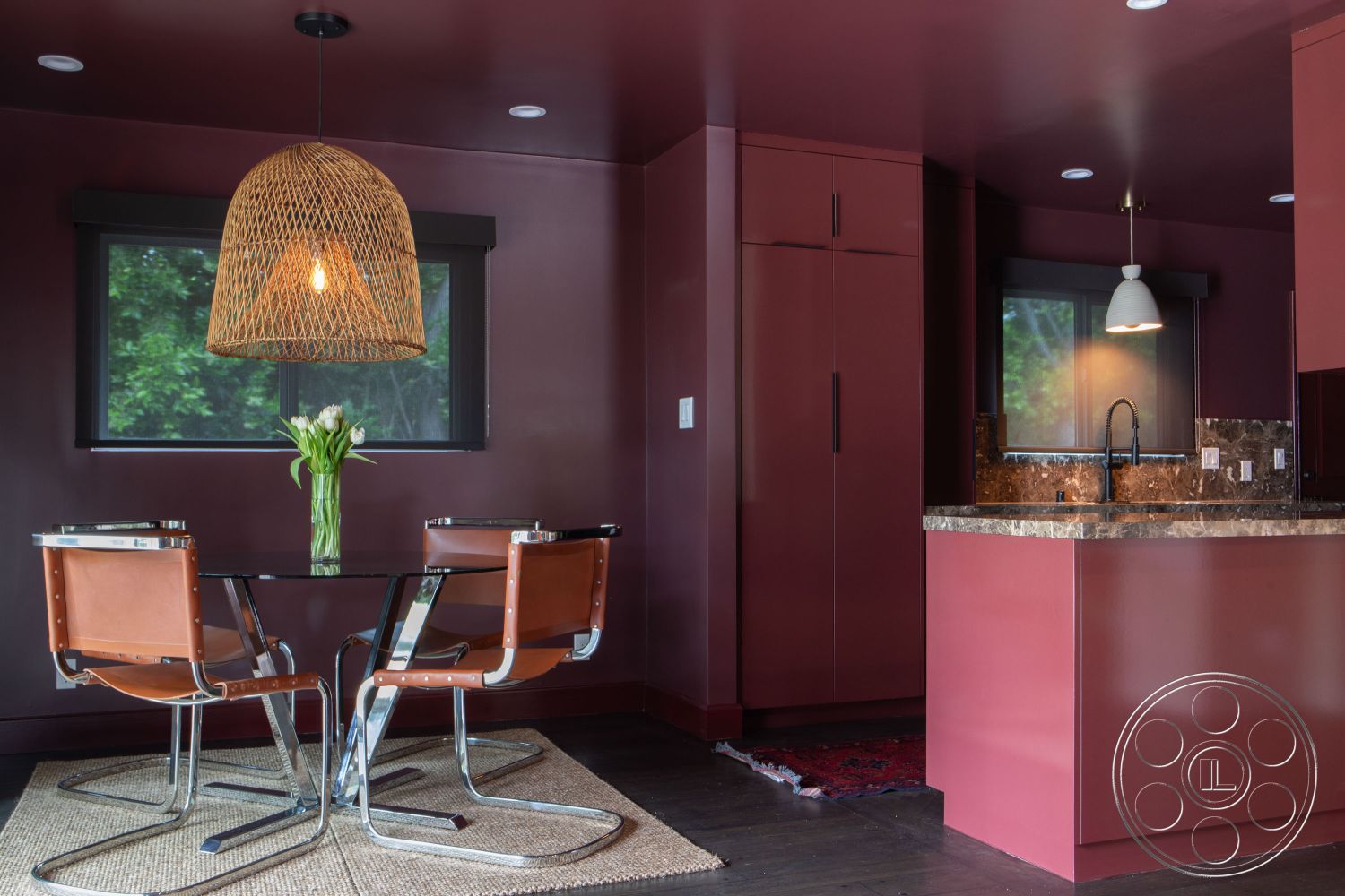 Bungalow 56 - dark hardwood flooring, woven area rug, modern minimalist decor, woven pendant light fixture, open kitchen layout, round glass dining table, interior kitchen with dark maroon walls, marble kitchen countertop, glass kitchen backsplash, wooden kitchen cabinets, chrome base dining chairs, floor-to-ceiling kitchen windows