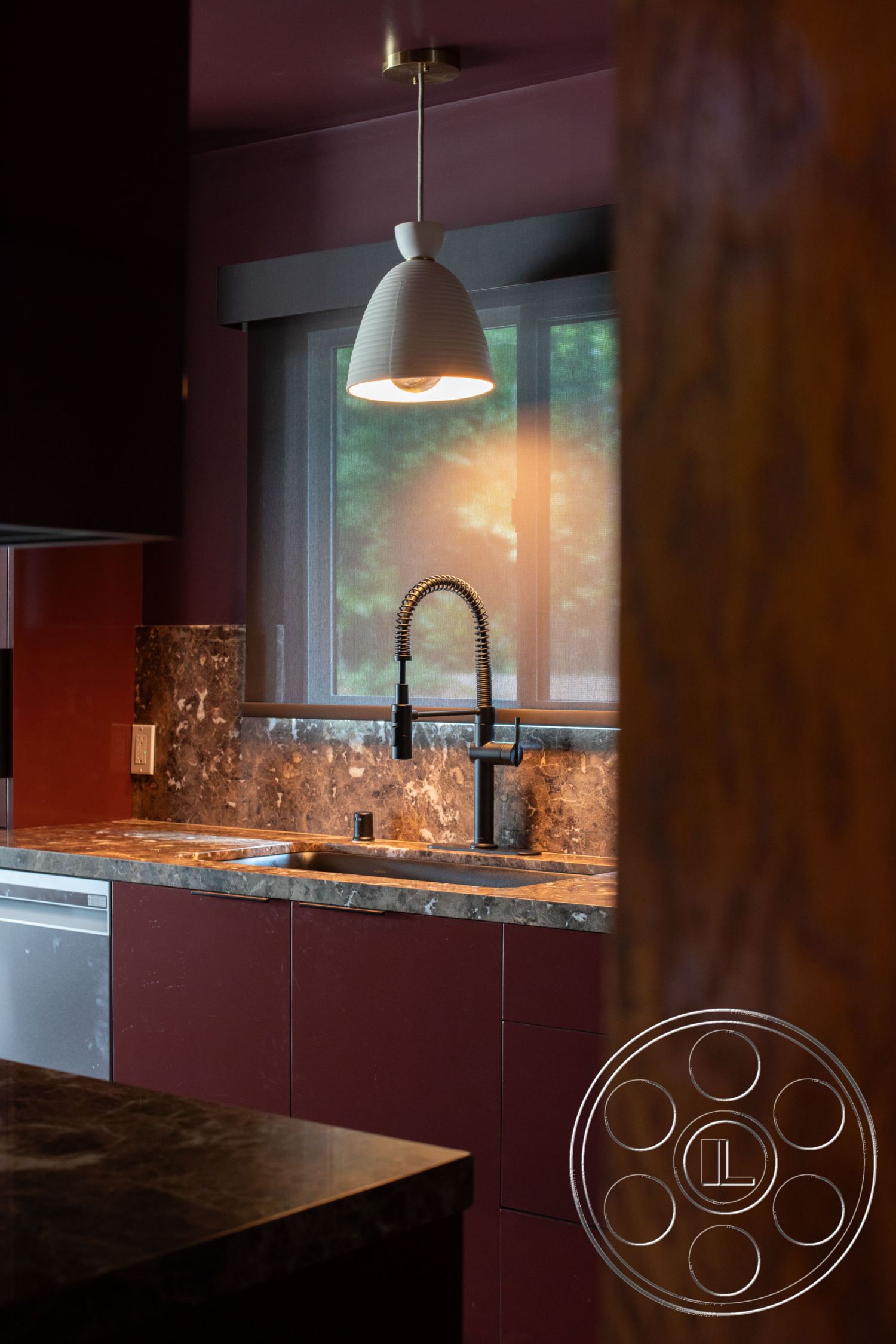 Bungalow 56 - stainless steel appliances, pendant light fixture, window with sheer curtains, contemporary kitchen design, black faucet with pull-down sprayer, marble kitchen countertop, interior kitchen with dark red cabinetry, textured wall treatment in muted burgundy, dark stone backsplash, modern cabinetry with clean lines