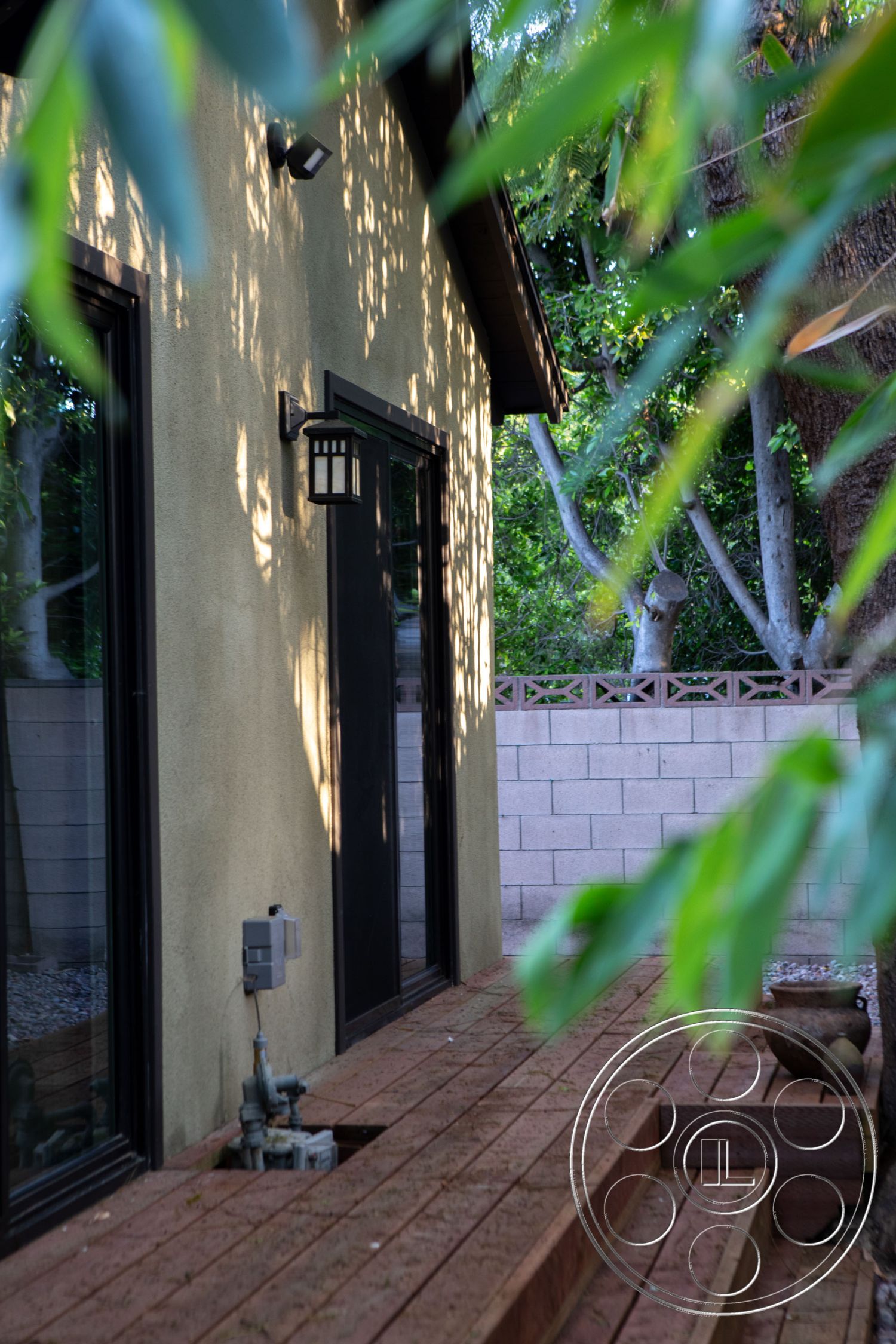 Bungalow 56 - modern architecture, minimalist design elements, black framed windows, suburban environment, outdoor lighting fixtures, residential home, landscaped garden setting, stucco exterior walls, wooden deck patio flooring, straight steps leading to entrance, exterior wall-mounted lantern, concrete block privacy wall, nature view surrounding property, exposed piping on patio, ground-level access