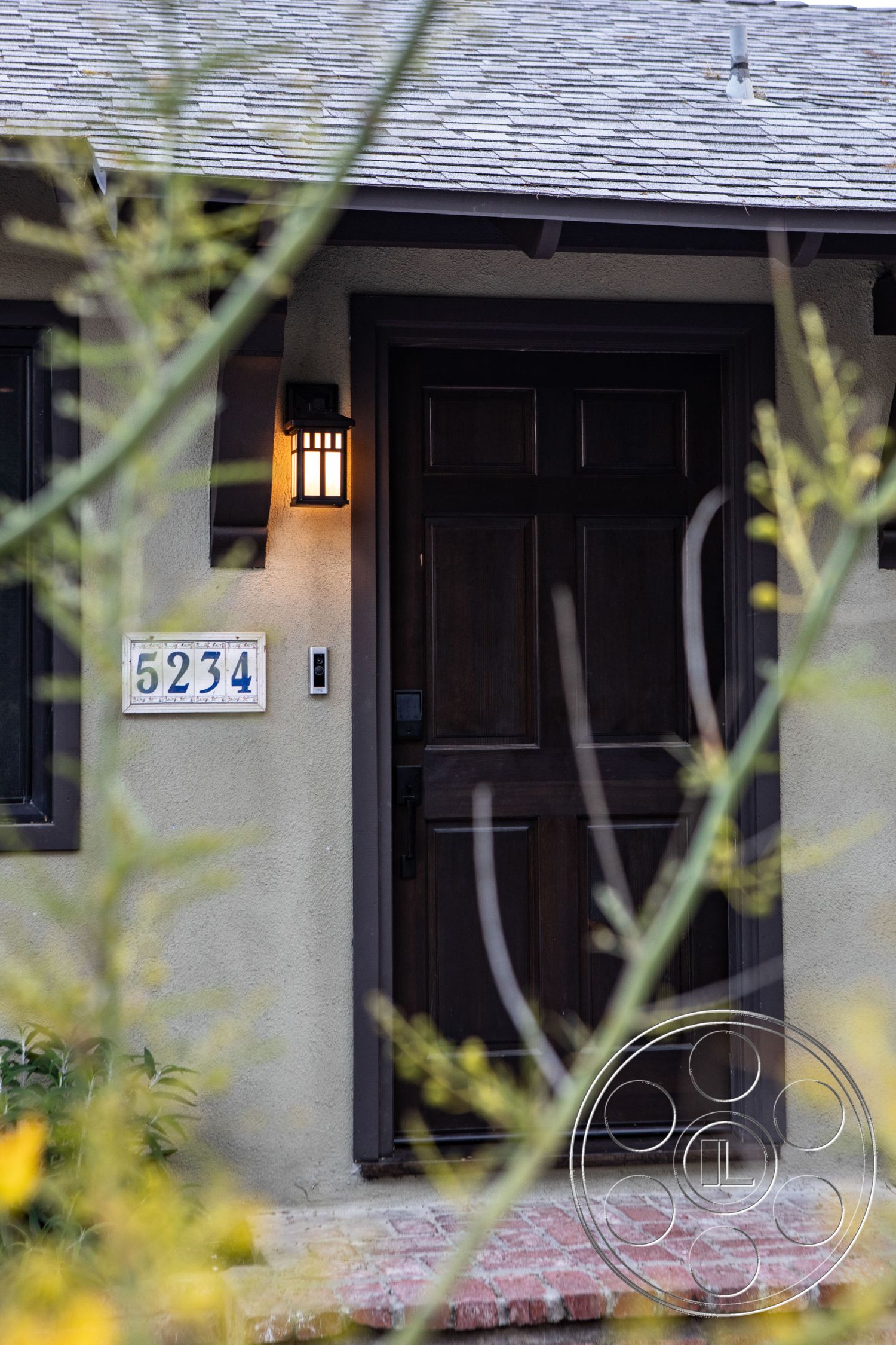 Bungalow 56 - exterior, brick pathway, stucco walls, craftsman architectural style, wood front door, shingle roof, lantern style wall light, decorative house numbers, landscaped front garden, natural plant elements