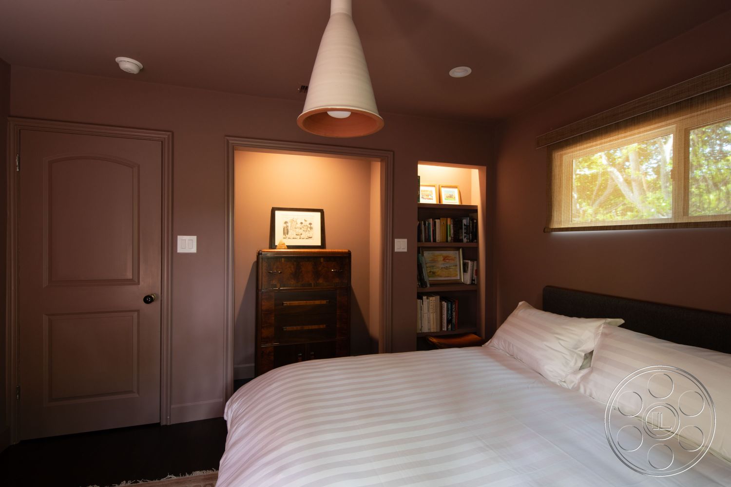 Bungalow 56 - cozy reading nook, window with bamboo shades, natural light from window, interior bedroom, dark hardwood bedroom floors, painted walls in warm brown, white bedding with striped pattern, minimalist pendant light fixture, wooden door with round black knob, built-in bookshelf with various books, decorative art on wall, doorway access to another room