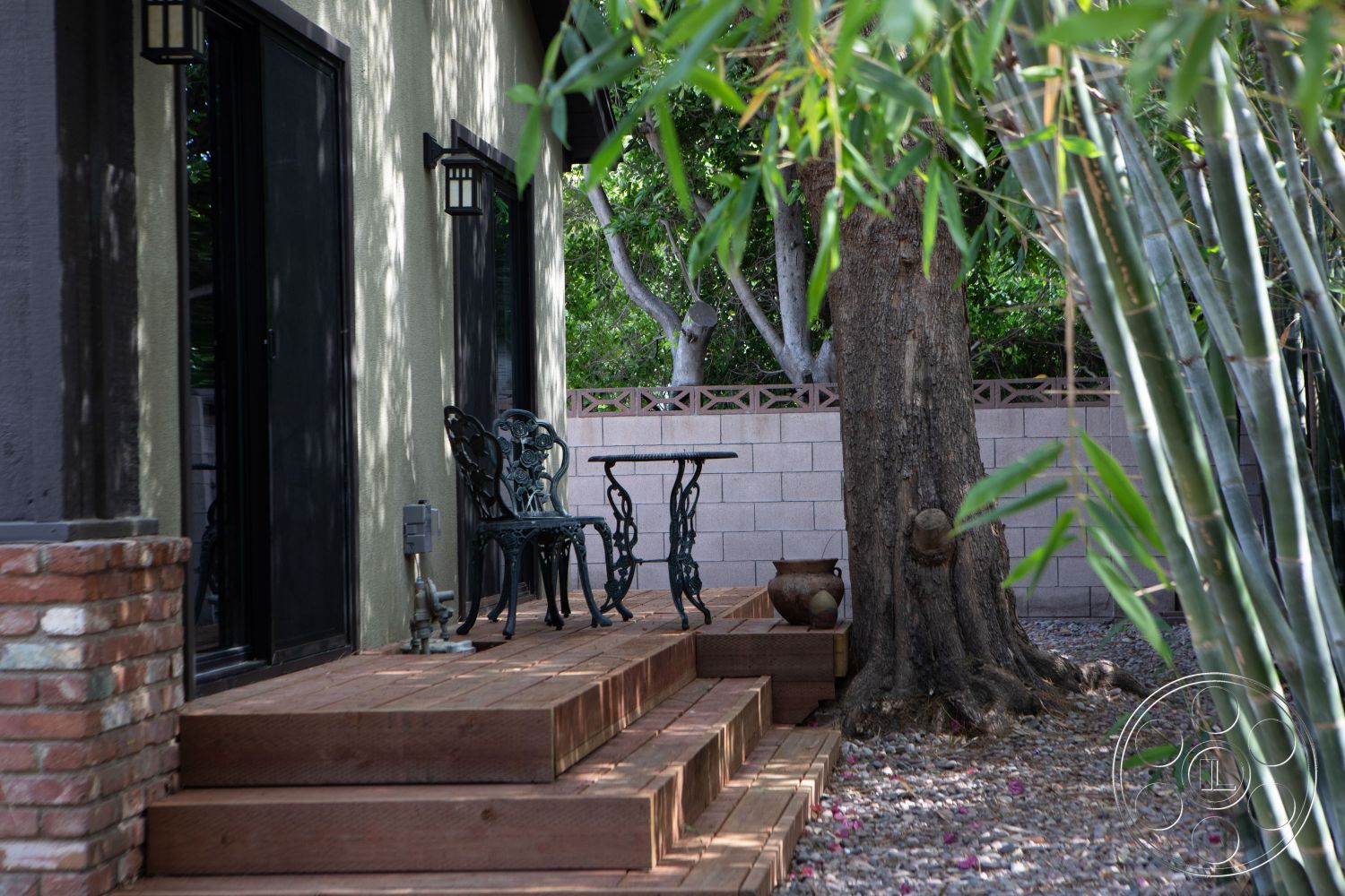 Bungalow 56 - outdoor seating area, interior-exterior flow, black framed sliding doors, natural stone planters, metal patio furniture, concrete block wall, contemporary garden design, landscaped garden setting, bamboo privacy screening, wood deck steps, wooden patio flooring, exposed brick column, mature tree landscaping, warm-toned wooden textures, rustic outdoor decor, shaded patio space