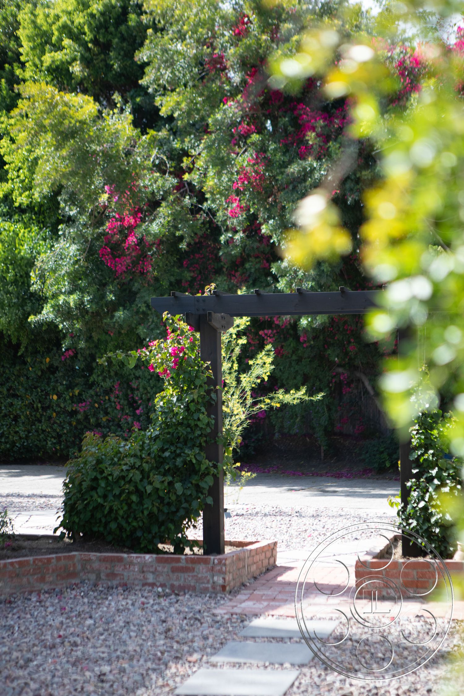 Bungalow 56 - landscape design, wooden pergola, colorful flowering plants, gravel driveway, suburban backyard, brick garden wall, exterior garden pathway, sandstone stepping stones, brick border flower bed, lush green shrubbery, bougainvillea flowering plants, decorative stone landscaping, natural stone mulch, pathway stone pavers, outdoor garden area, low-maintenance horticulture