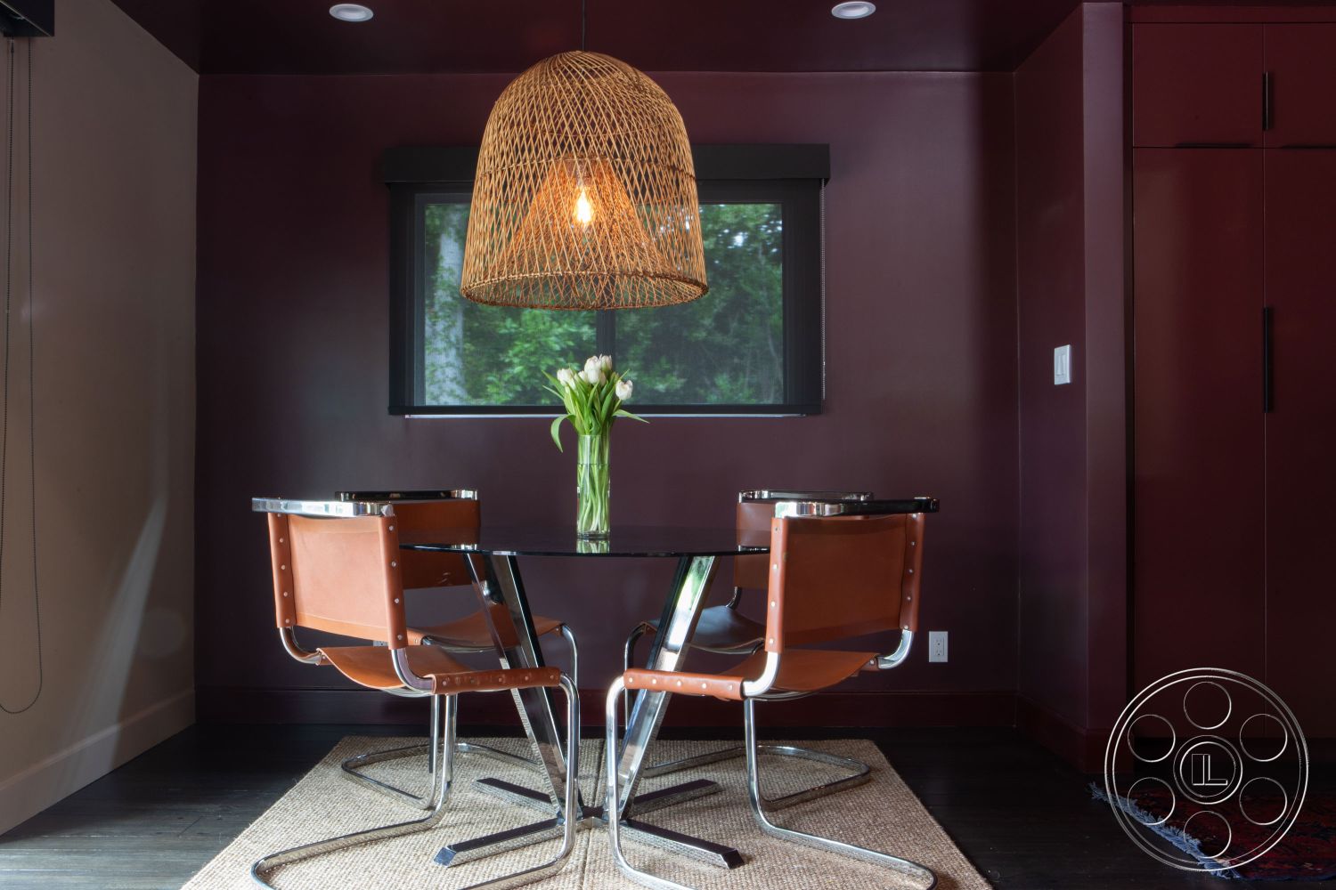 Bungalow 56 - textured area rug, glass dining table, dark wood flooring, modern minimalist decor, interior dining area, woven pendant light fixture, burgundy painted walls, metal frame dining chairs, leather seat dining chairs, large window with forest view, natural light-filled space