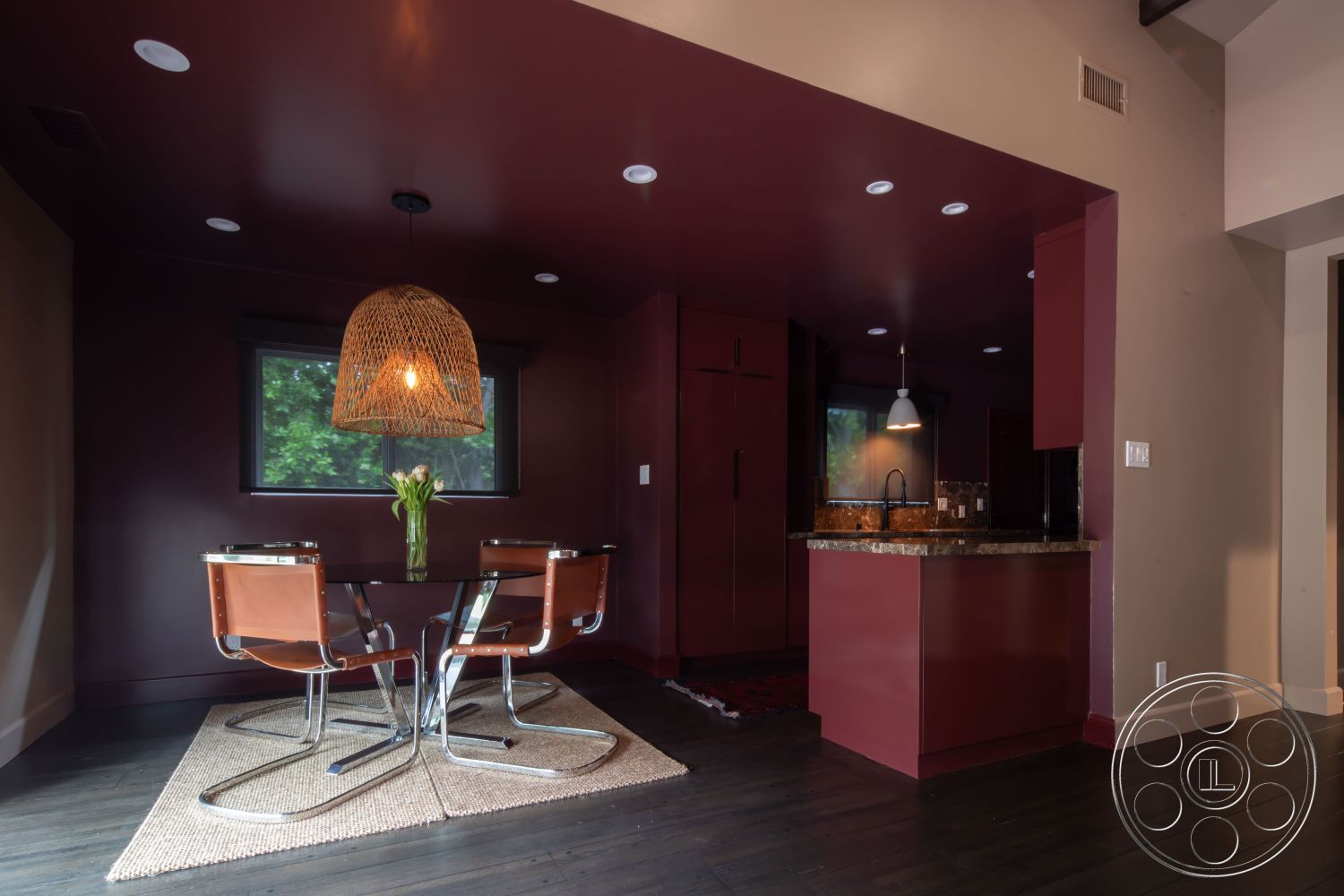 Bungalow 56 - hardwood flooring, textured area rug, floor-to-ceiling windows, woven pendant light fixture, open concept dining area, round glass dining table, dark maroon painted walls, minimalist kitchen design, black granite kitchen countertop, modern chrome dining chairs, natural light filling the space, contemporary interior style, small breakfast nook, decorative vase with fresh flowers