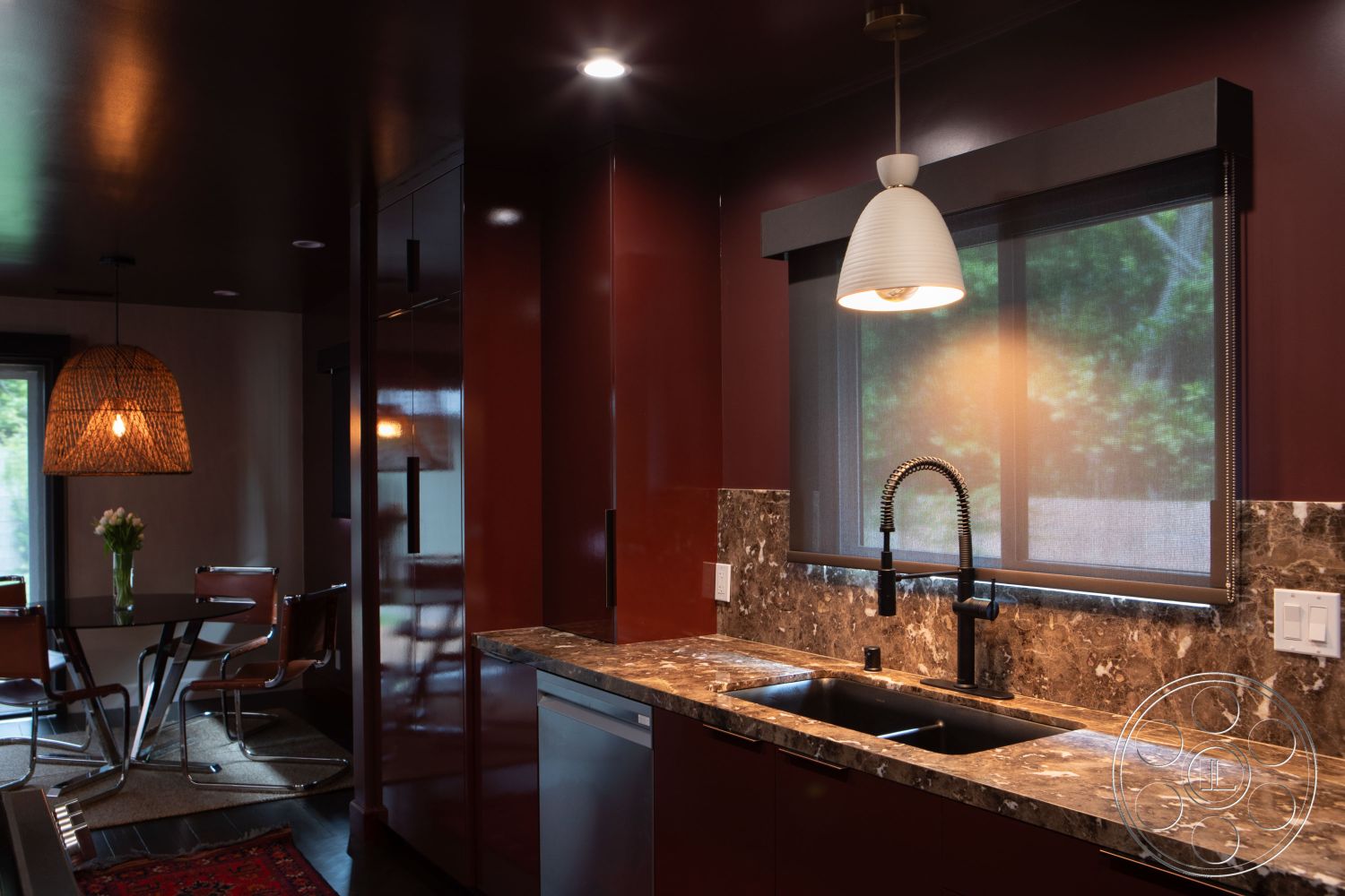 Bungalow 56 - textured wall treatment, woven pendant light fixture, contemporary kitchen design, marble kitchen countertop, interior kitchen with dark red cabinetry, stainless steel sink with modern faucet, glass kitchen window with roller shades, open dining area with round glass table, metal dining chairs with leather accents, black hardwood flooring