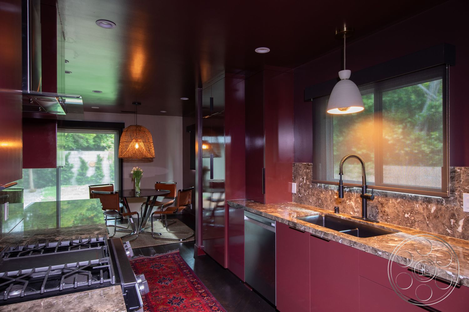 Bungalow 56 - stainless steel appliances, dark wood flooring, natural light from large windows, open-concept layout, black marble countertops, integrated cabinetry, modern dining area, round glass dining table, interior kitchen with burgundy cabinetry, pendant lighting fixture, minimalist kitchen design, brown leather dining chairs, glass cooktop, textured ceiling treatment, glass sliding door to outdoor space, wicker pendant light, decorative area rug with intricate patterns, cozy breakfast nook