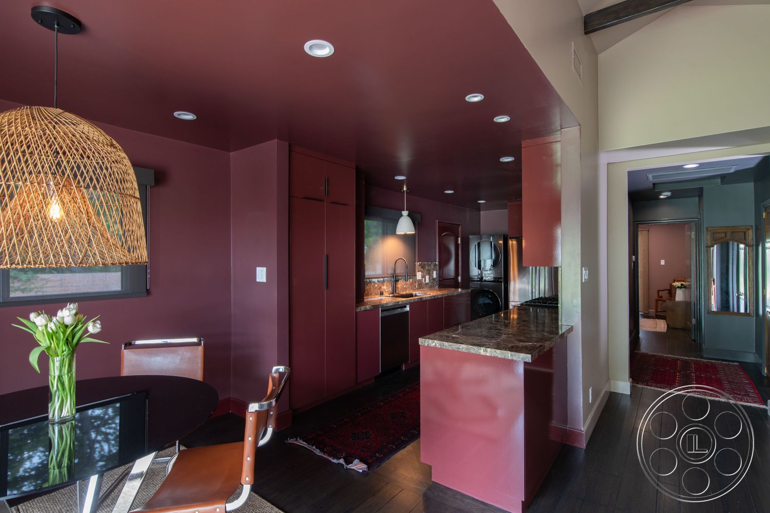 Bungalow 56 - neutral color palette, decorative area rug, stainless steel appliances, round black dining table, contemporary aesthetic, textured wall treatments, warm accent lighting, modern kitchen design, woven pendant light fixture, interior kitchen with dark wood floors, glossy burgundy cabinets, granite kitchen countertops, open floor plan layout, glass backsplash behind the sink, cozy dining nook with natural light, contrast between dark and light colors, spacious living area with adjacent hallway, minimalist furniture style, high ceilings with exposed wood beams