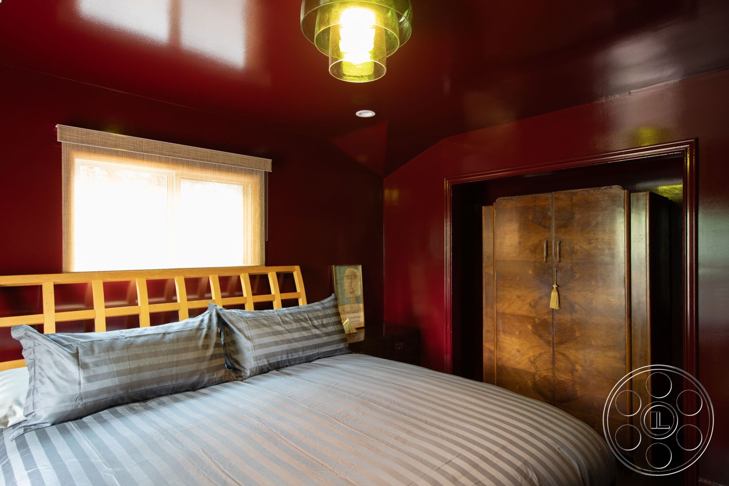 Bungalow 56 - warm color palette, contemporary design elements, wooden bed frame, vintage wooden wardrobe, interior bedroom space, high-gloss maroon wall finish, striped gray bedding, decorative wooden headboard, textured ceiling detail, large double-pane window, natural light illumination, flush-mounted pendant lighting, smooth wood wardrobe surface, minimalist decor accents, functional bedroom layout