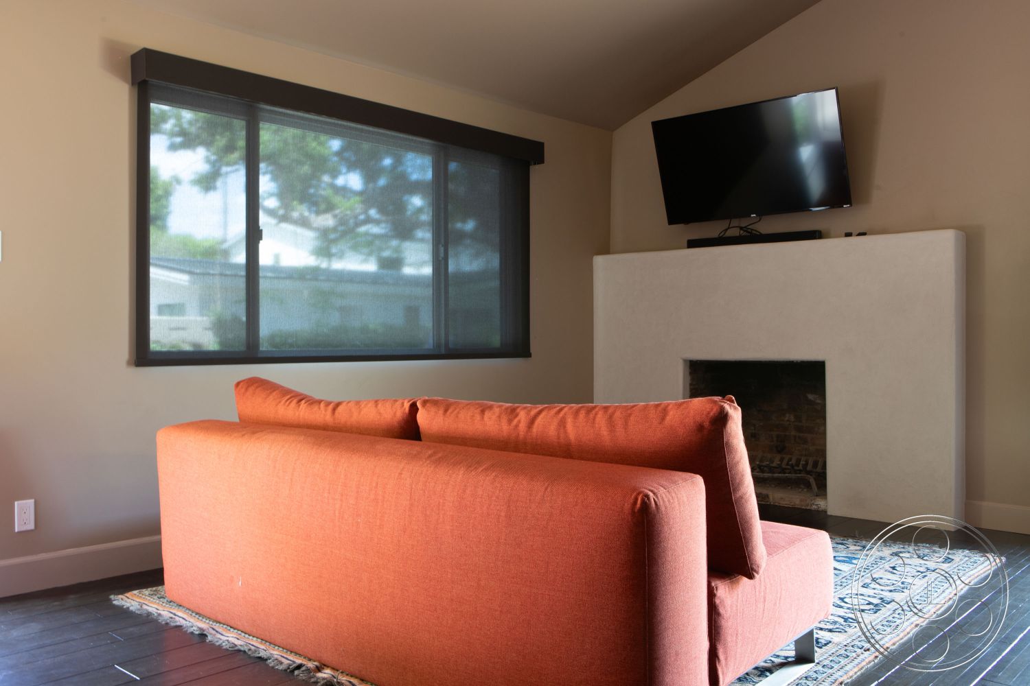 Bungalow 56 - minimalist decor, neutral wall colors, open floor plan, dark wood flooring, contemporary design elements, natural light from windows, textured wall treatment, interior living room, orange upholstered sofa, white plaster fireplace, black mounted TV, large windows with dark frames, area rug with intricate pattern