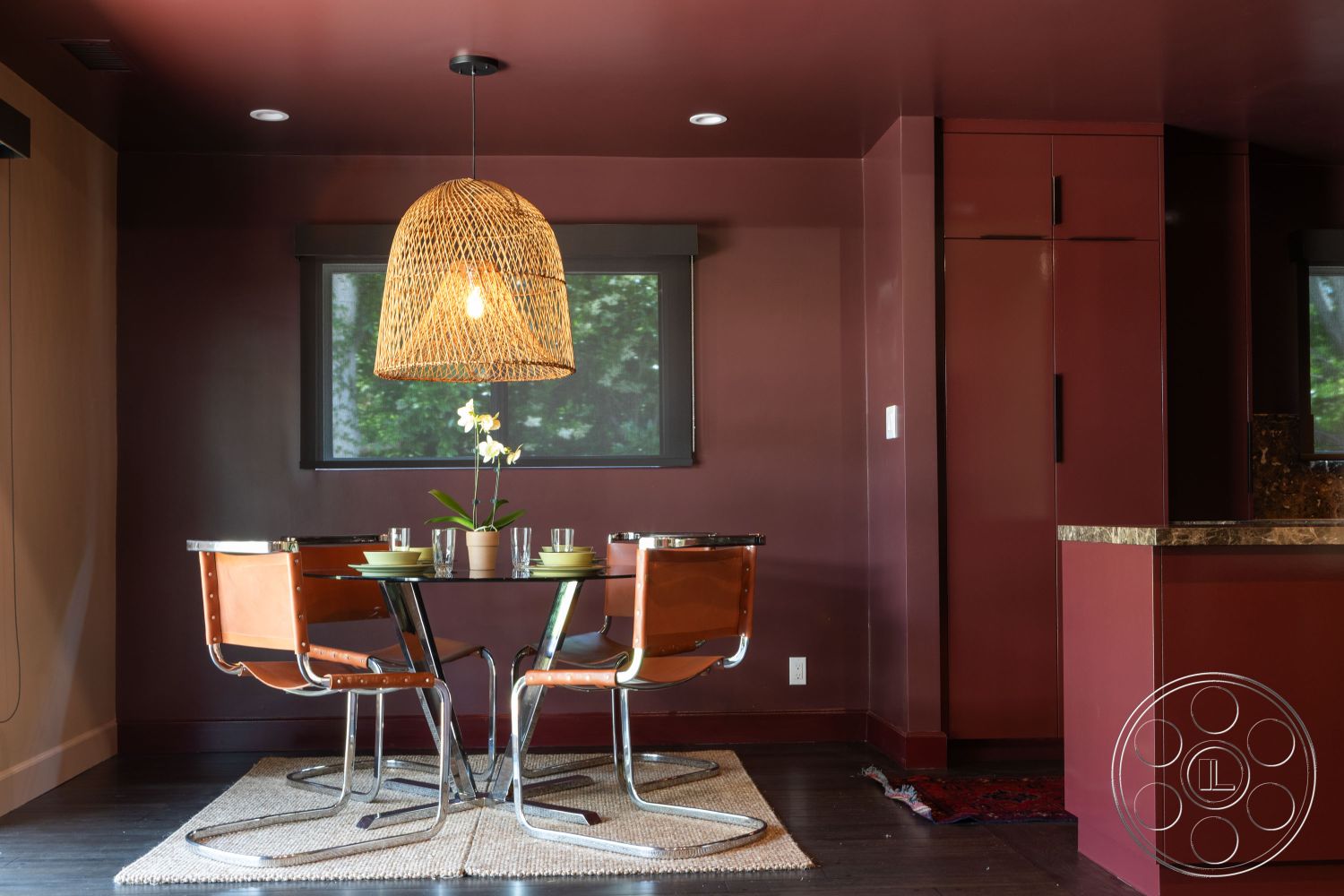 Bungalow 56 - minimalist decor, textured area rug, floor-to-ceiling windows, interior dining area, woven pendant light fixture, granite kitchen countertop, open kitchen layout, contemporary lighting design, dark red wall paint, wooden floorboards, modern round dining table, metal frame chairs with leather seats, built-in storage cabinets, greenery centerpiece