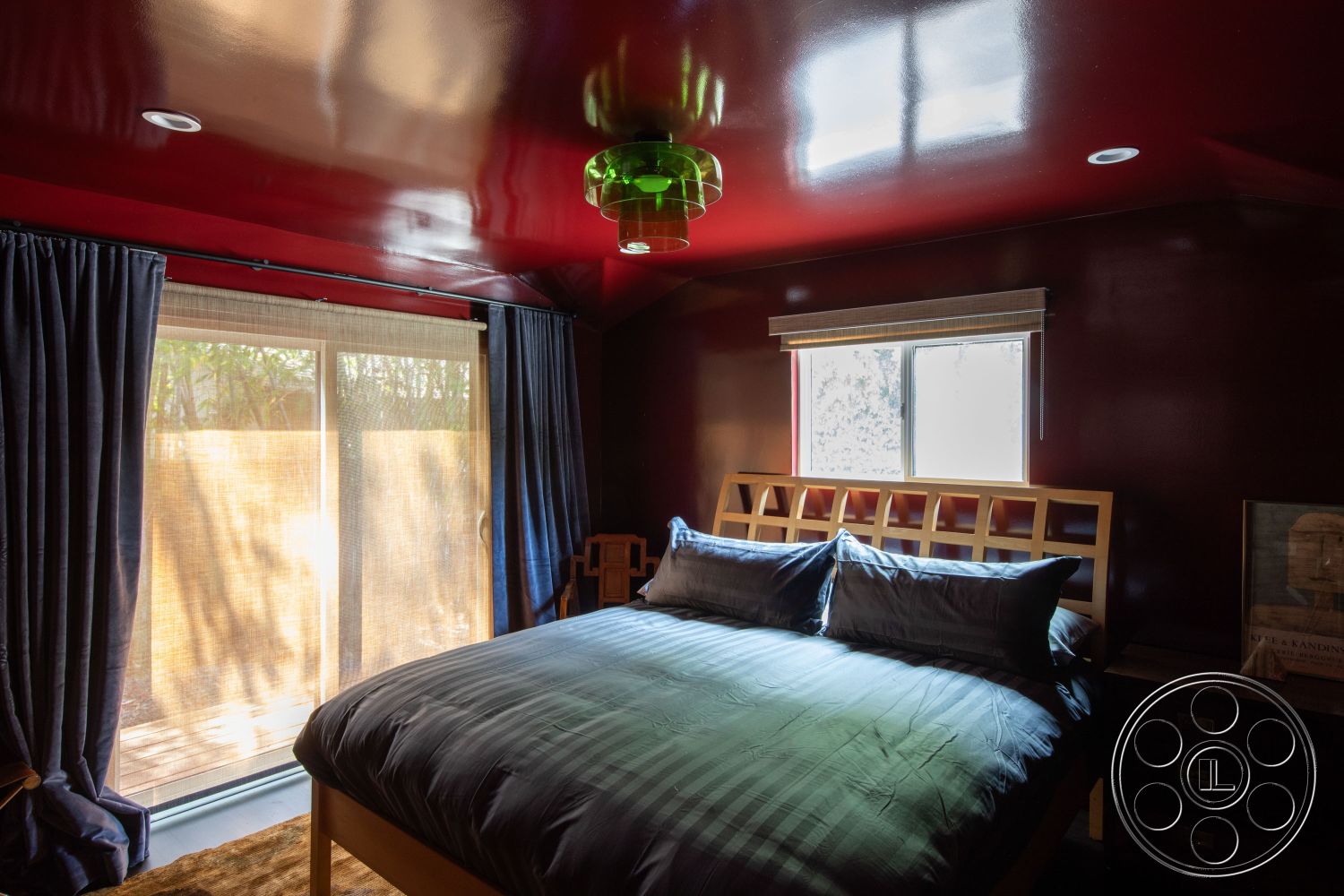 Bungalow 56 - cozy atmosphere, natural light from window, interior bedroom, red high-gloss painted walls, vaulted ceiling with gloss finish, contemporary wooden bed frame, soft blue bedding, sheer window shades, dark blue curtain drapes, sliding glass door to outdoor area, decorative floor lamp, textured carpet flooring, minimalist bedside table, modern green pendant light, wood frame window, geometric headboard design