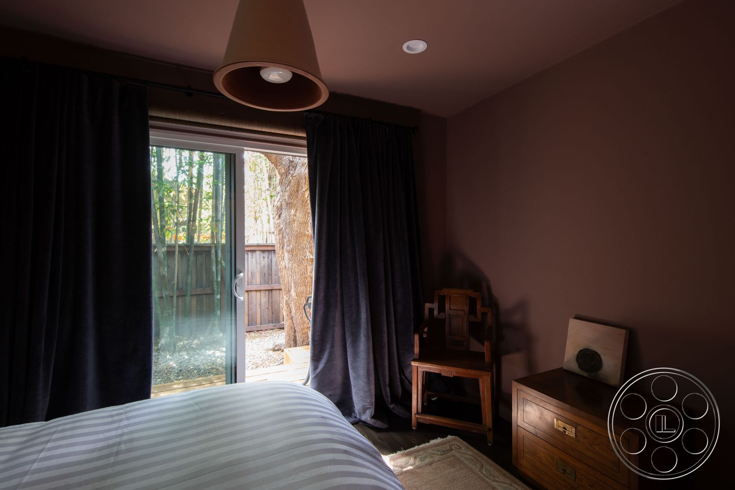 Bungalow 56 - interior bedroom with dark wood paneling, textured plaster walls in warm tones, sliding glass door leading to outdoor space, floor-to-ceiling windows with natural light, blackout curtains in soft fabric, hardwood bedroom floors with a rich finish, minimalist wooden accent chair, decorative wall art in neutral colors, contemporary bedside table with metallic accents, cozy atmosphere with soft lighting, patio access for indoor-outdoor living