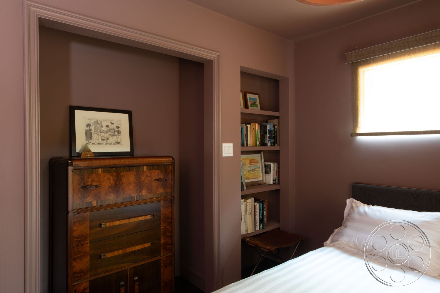 Bungalow 56 - exposed wood ceiling beams interior, purple painted walls interior, wooden dresser with decorative inlays interior, built-in bookcase with books interior, corner window with soft roman shades interior, textured fabric bed linens interior, modern minimalist nightstand interior, framed artwork on wall interior, natural light from window interior, curved wall edge detail interior