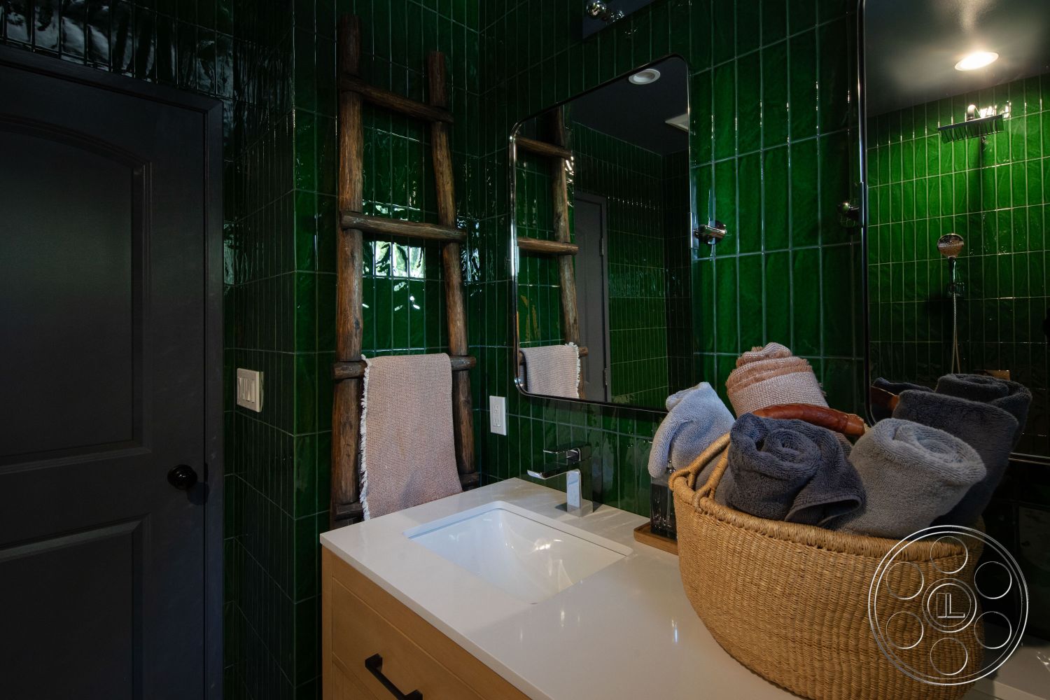 Bungalow 56 - neutral color palette, spa-like atmosphere, minimalist design elements, textured wall surface, geometric tile pattern, white quartz countertop vanity, interior bathroom with green glazed tile walls, natural wood ladder towel rack, modern black faucet fixtures, dark gray painted door, large mirror with wall-mounted lighting, woven basket for towels, hygienic wall-mounted soap dispenser, high-gloss ceramic tile backsplash, natural light from shower area, functional storage solutions