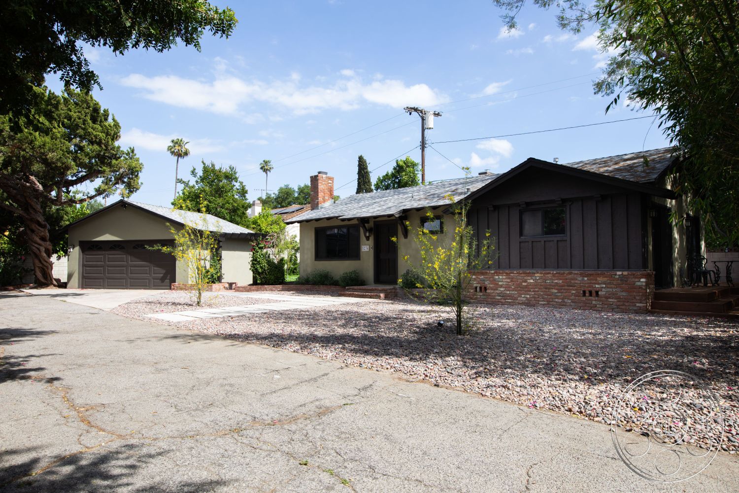 Bungalow 56 - natural light, asphalt shingle roof, low-maintenance landscaping, lush greenery, landscaped front yard, brick accent wall, concrete driveway, wood siding, exterior house facade, driveway gravel surface, single-story ranch-style home, detached garage, patio gravel flooring, wooden porch steps, exposed chimney, neighborhood setting with trees, traditional home design, polished concrete walkways, solar panel potential