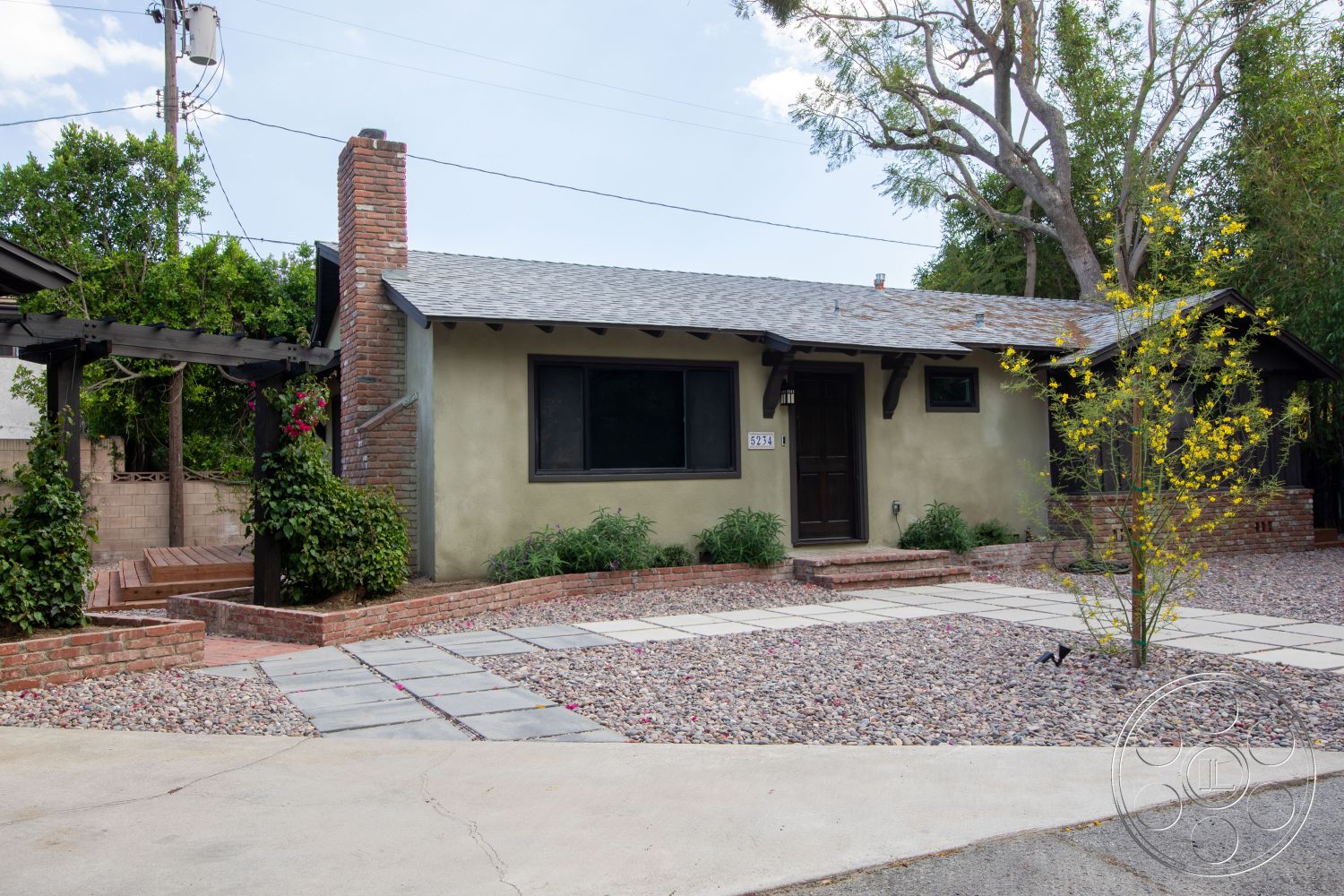 Bungalow 56 - exterior, wooden front door, brick chimney, flagstone walkway, stucco exterior walls, gabled asphalt shingle roof, pergola with wood beams, gravel landscape bed, brick patio border, small shrubs landscaping, open front porch, city suburban setting