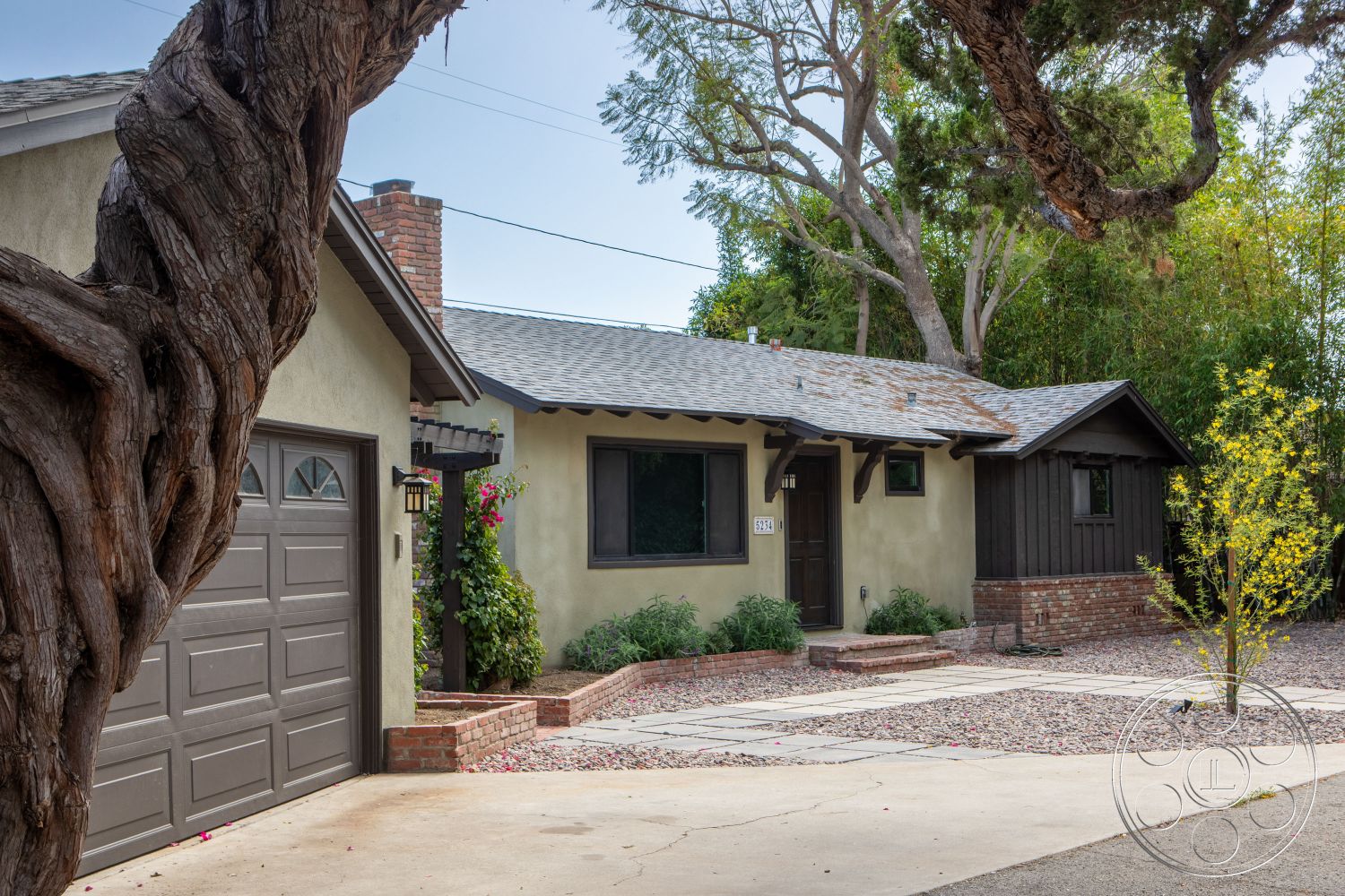 Bungalow 56 - asphalt shingle roof, single-story home, natural stone pathway, craftsman architectural style, horizontal wood siding, stucco exterior walls, landscaped front garden, exposed brick chimney, dark wood garage door, brick front steps, pebble driveway, wooden porch overhang, window planters with flowers, textured stucco finish, leafy tree canopy, suburban front yard setting