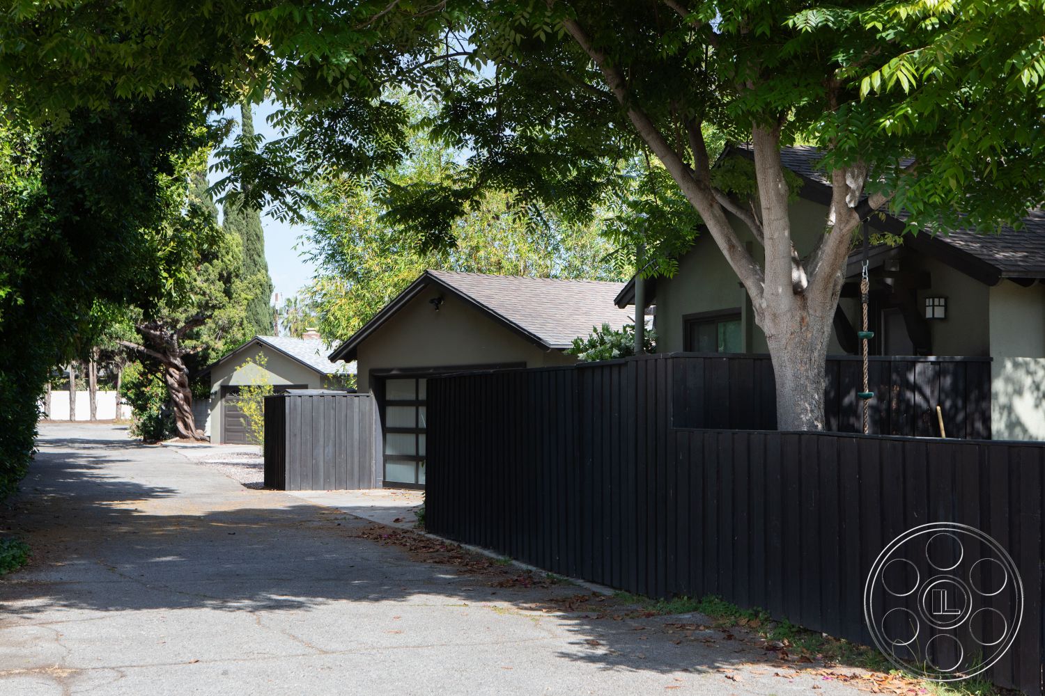Bungalow 56 - mature trees, single-story home, paved pathway, gabled roof, asphalt driveway, suburban backyard, landscaped garden area, sloped roof design, wood fence panels, exterior home facade, grey stucco walls, attached garage, residential alleyway, nautral stone accent landscaping