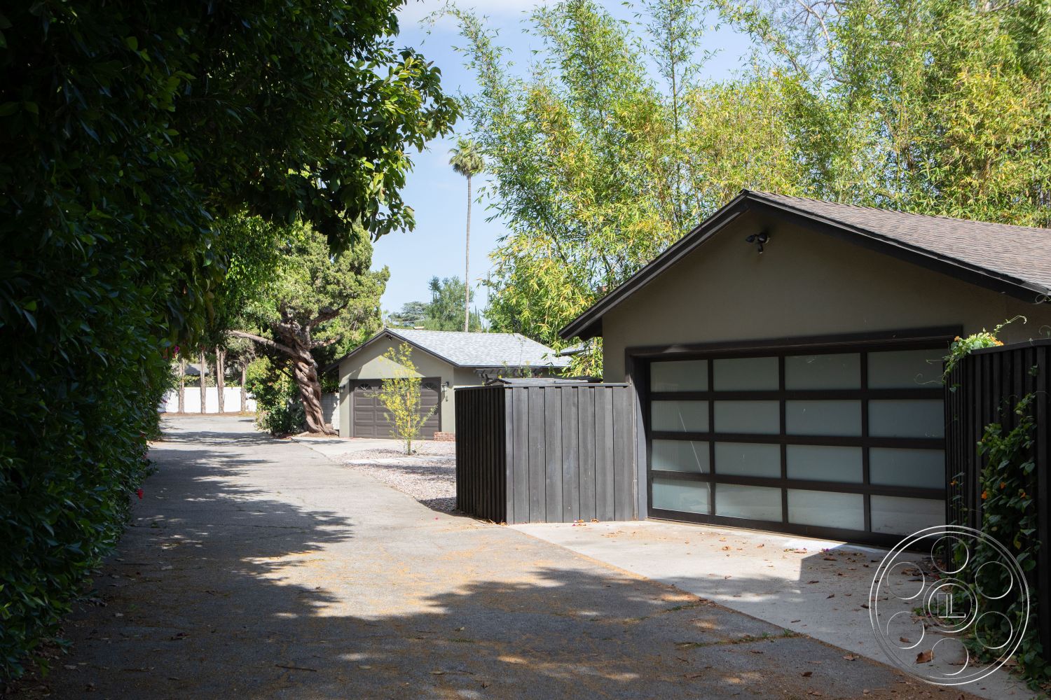 Bungalow 56 - natural light setting, exterior garage with frosted glass doors, asphalt driveway leading to the garage, concrete driveway surface, landscape with mature trees, bamboo border along the pathway, wooden fence with dark finish, rhythm of contemporary architectural lines, single-story garage structure, residential setting with greenery, modern home aesthetic, neighborhood with a suburban character, open air circulation, casual outdoor pathway