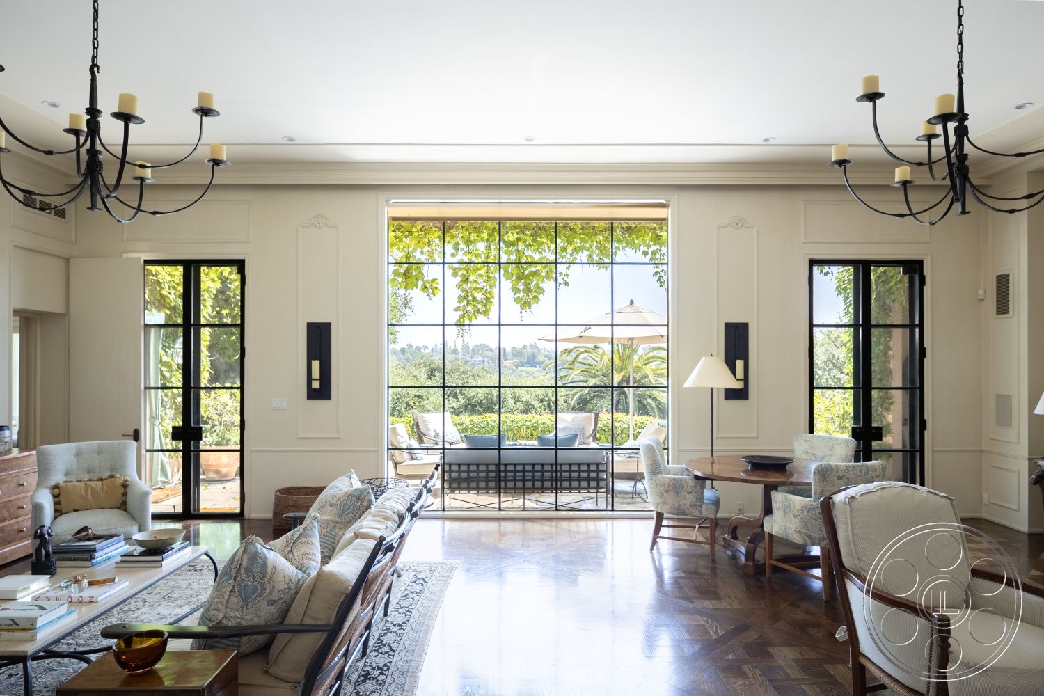 The Regent - landscaped garden view, large floor-to-ceiling windows, decorative wall paneling, exposed wood ceiling beams, white painted walls, traditional chandelier lighting, parquet hardwood living room floors, decorative crown molding details, modern glass sliding doors, french doors opening to garden patio, black steel door frames, comfortable upholstered seating, stylish coffee table arrangement, textured area rug with intricate patterns, elegant dining table setup, traditional armchair with blue fabric, sunlit room with natural light, open layout living space, contemporary home decor accents, lush greenery surrounding the property