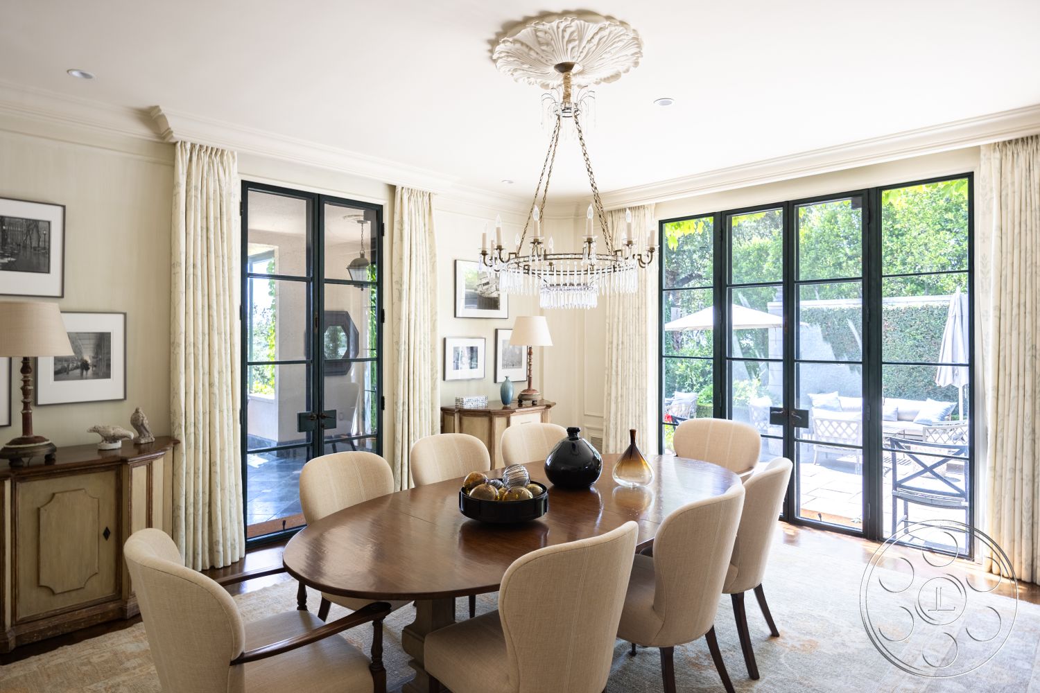 The Regent - glass sliding doors, textured plaster walls, photographic wall art, round wooden dining table, curtained windows, chandelier with crystal accents, hardwood dining room floors, cream upholstered dining chairs, black steel frame doors, exposed wood crown molding, decorative ceiling medallion, neutral color curtains, dining room with garden view, transitional interior style, wood sideboard cabinet, outdoor patio area