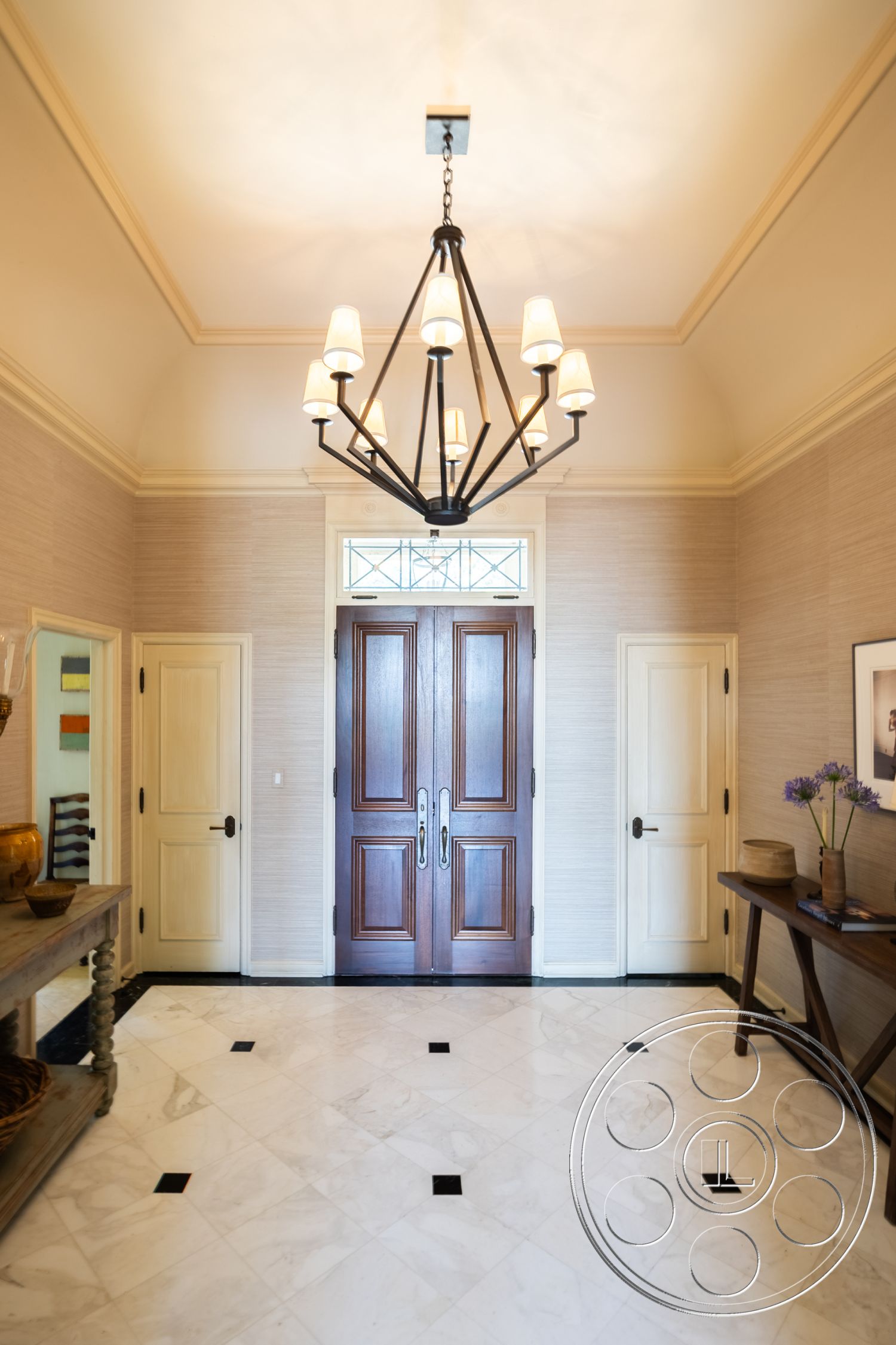 The Regent - neutral color palette, light-filled foyer, interior entryway, marble flooring with black accents, exposed wood ceiling details, traditional double front doors, brushed brass chandelier, textured wallpaper accent, cream-colored wall panels, dark wood console table, elegant decor vase, traditional molding details, welcoming entry space, stylish door hardware, cohesive design elements