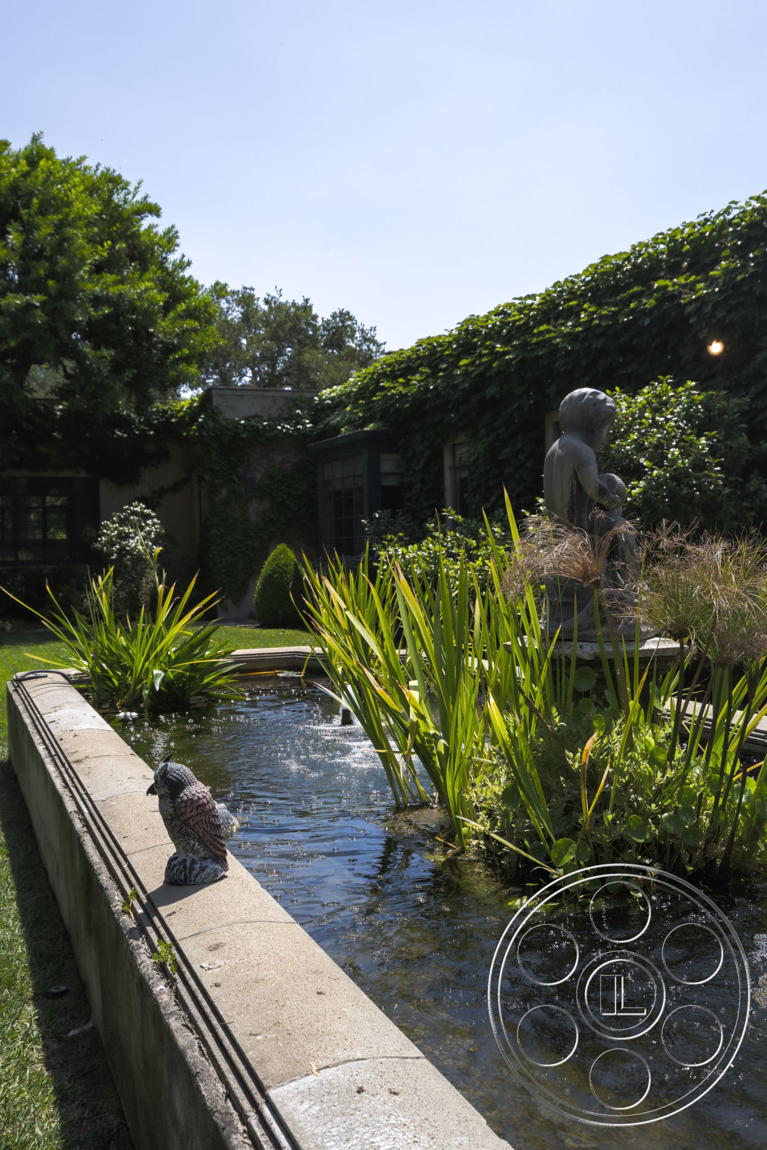 The Regent - natural light, vibrant green foliage, garden patio, ornamental grasses, landscaped garden area, suburban garden, exterior stone water feature, stone border around pond, lush garden surroundings, sculptural garden statue, concrete pond edge, clear blue sky view, serene outdoor setting, mixed plantings, decorative bird statue