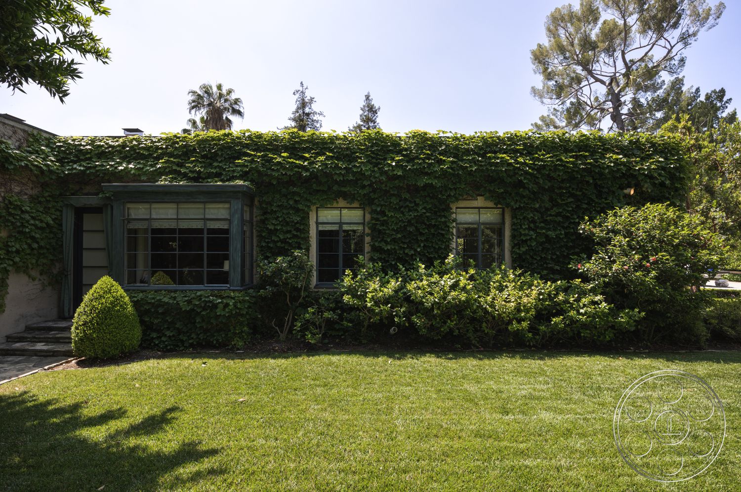 The Regent - manicured lawn, suburban backyard, low hedges, outdoor lighting fixtures, dormer windows, wood window frames, sunny exterior, landscaped garden setting, modern ranch-style home, exterior ivy-covered walls, curved pathway with stone pavers, greenery around the house, tall trees in the vicinity, casual garden patio