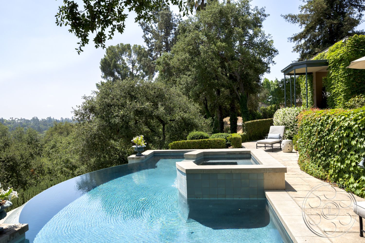The Regent - curved pool edge, infinity pool with tile coping, rectangular spa with concrete surround, natural stone patio flooring, landscape garden with hedges and mature trees, outdoor lounge area with modern chairs, lush green hedge privacy, wooden pergola structure, city skyline view in the background, exterior pool area, residential backyard oasis, stone planter with flowering plants, textured walls of surrounding foliage
