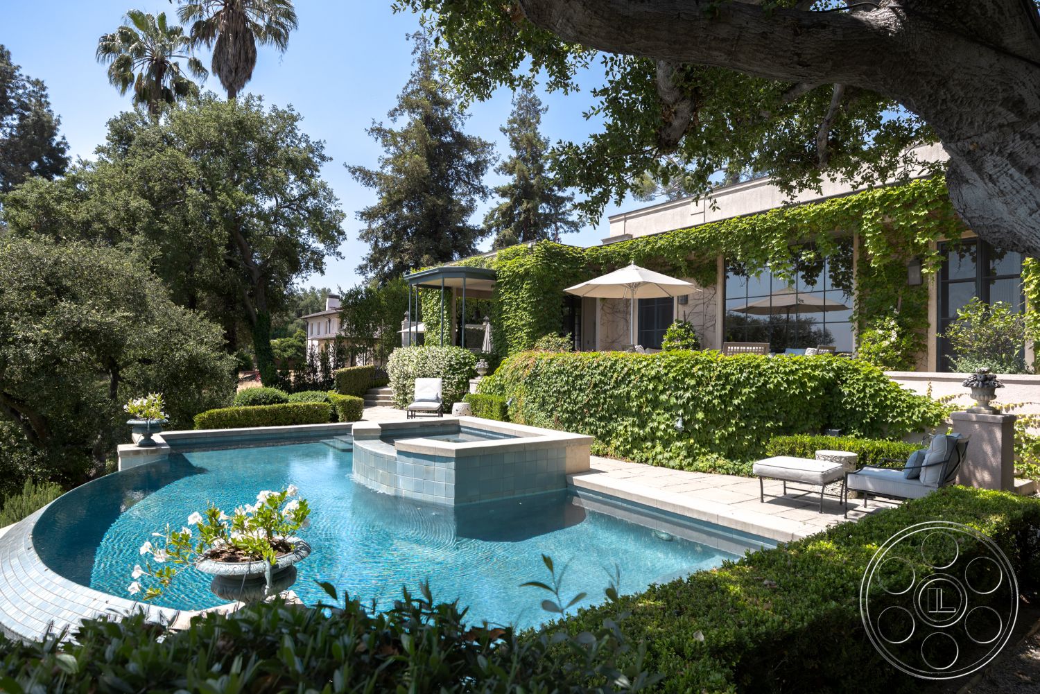 The Regent - exterior, outdoor seating area, glass windows, tropical palm trees, rectangular pool, stone pool deck, concrete patio floor, blue glass tile pool finish, nature landscaping with trees, stucco home exterior, hardscaped terrace area, in-ground swimming pool feature, metal pergola with shade, garden hedge borders, potted plants around pool area, irregularly shaped pool edges, natural stone steps leading to house, surrounding green foliage, elevated backyard setting