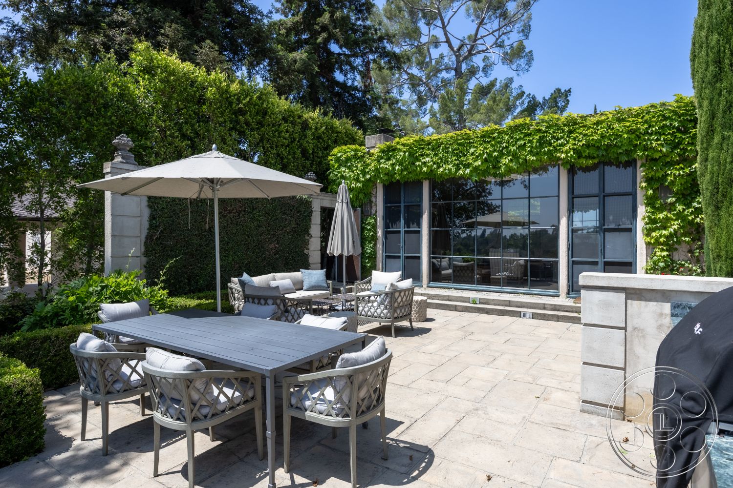 The Regent - seasonal foliage, modern outdoor furniture, surrounding greenery, exposed concrete wall, glass wall, cylindrical columns, metal outdoor chairs, sunny exterior, urban garden setting, rectangular patio layout, stone patio flooring, concrete wall planters, wooden outdoor dining table, large fabric umbrella, lush landscaping with hedges, aluminum railings