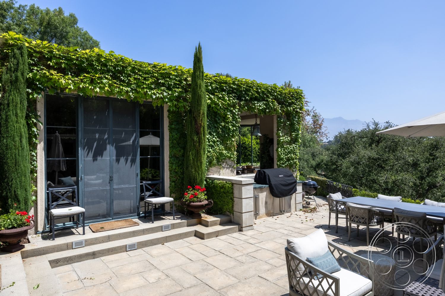 The Regent - landscaped garden patio, modern outdoor dining area, exterior patio flooring, flagstone outdoor flooring, stone masonry kitchen barbecue, aluminum outdoor dining furniture, sliding glass door entry, cascading ivy wall treatment, concrete steps with stone border, vegetation surround with cypress trees, mountain view terrace setting, open-air outdoor kitchen design, sunshade patio umbrella, rustic stone feature wall, decorative ceramic planters
