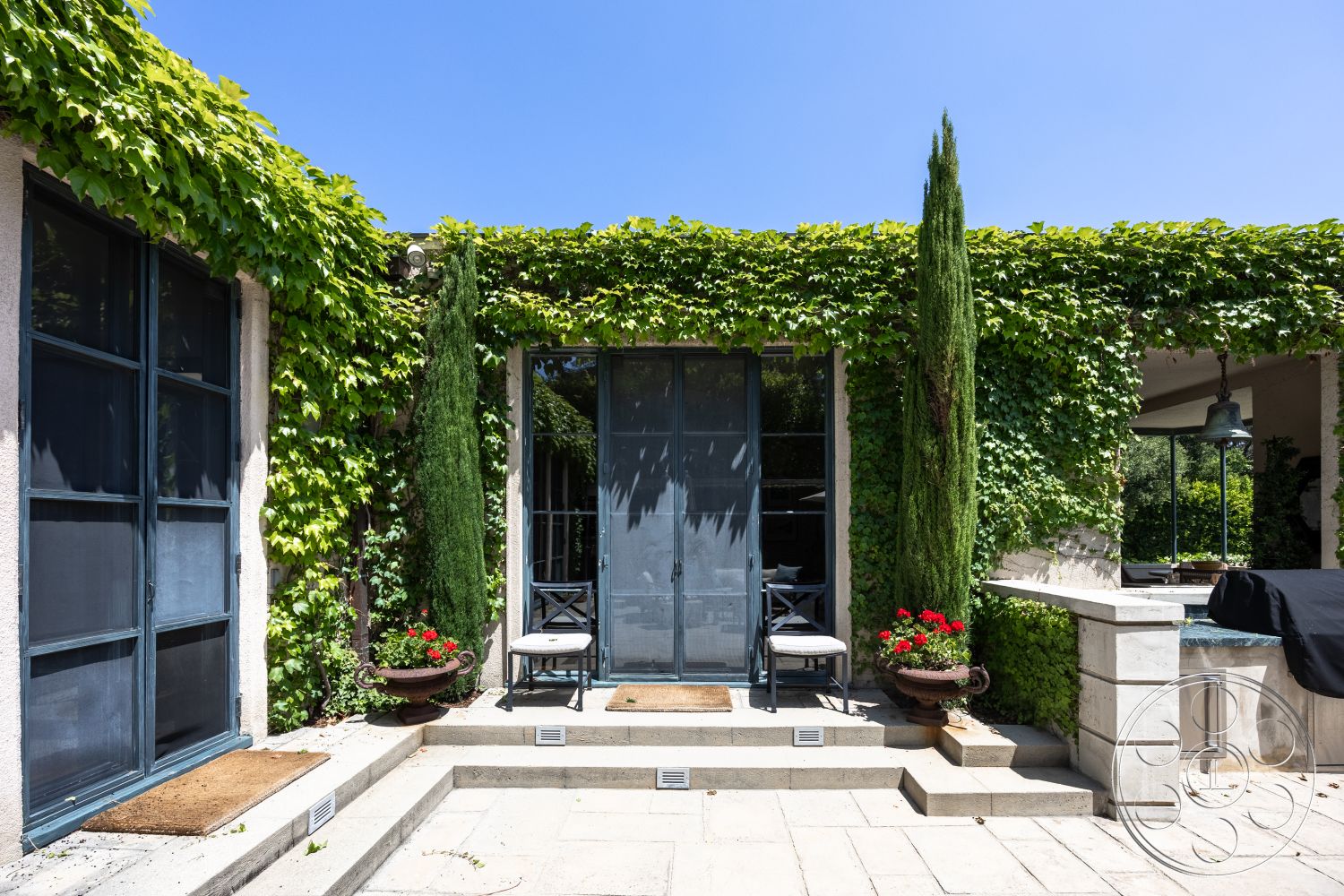 The Regent - natural landscaping, concrete steps, tall cypress trees, exposed stone wall, urban garden setting, stone patio flooring, decorative stone border, glass door entrance, vines on exterior walls, wooden outdoor chairs, potted flowers on patio, interior space with outdoor access, modern outdoor living area, pastel-painted walls, soft sunlight ambiance