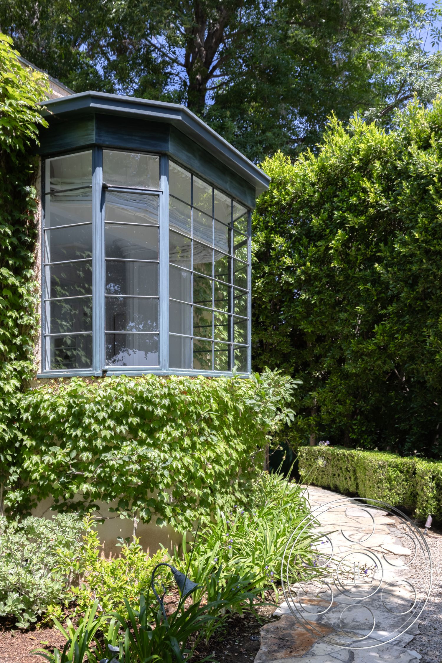 The Regent - exterior, low-maintenance landscaping, stone pathway, planted shrubs, urban garden, ivy-covered wall, landscaped garden beds, bay window with glass panes, curved bay window, natural stone exterior, greenery accent wall, leafy vine trellis, framed glass windows, stately garden setting, textured stone pavers, natural light infusion