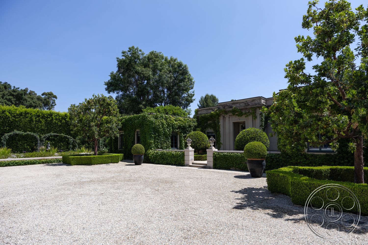 The Regent - exterior, gravel driveway, ornamental trees, boxwood hedges, formal garden layout, classic architectural details, symmetrical garden design, stone planters, topiary shrubs, lush green landscaping, elevated entryway, pergola with climbing vines, sunlit courtyard, modern villa exterior, private estate setting, rich foliage, minimalist garden elements