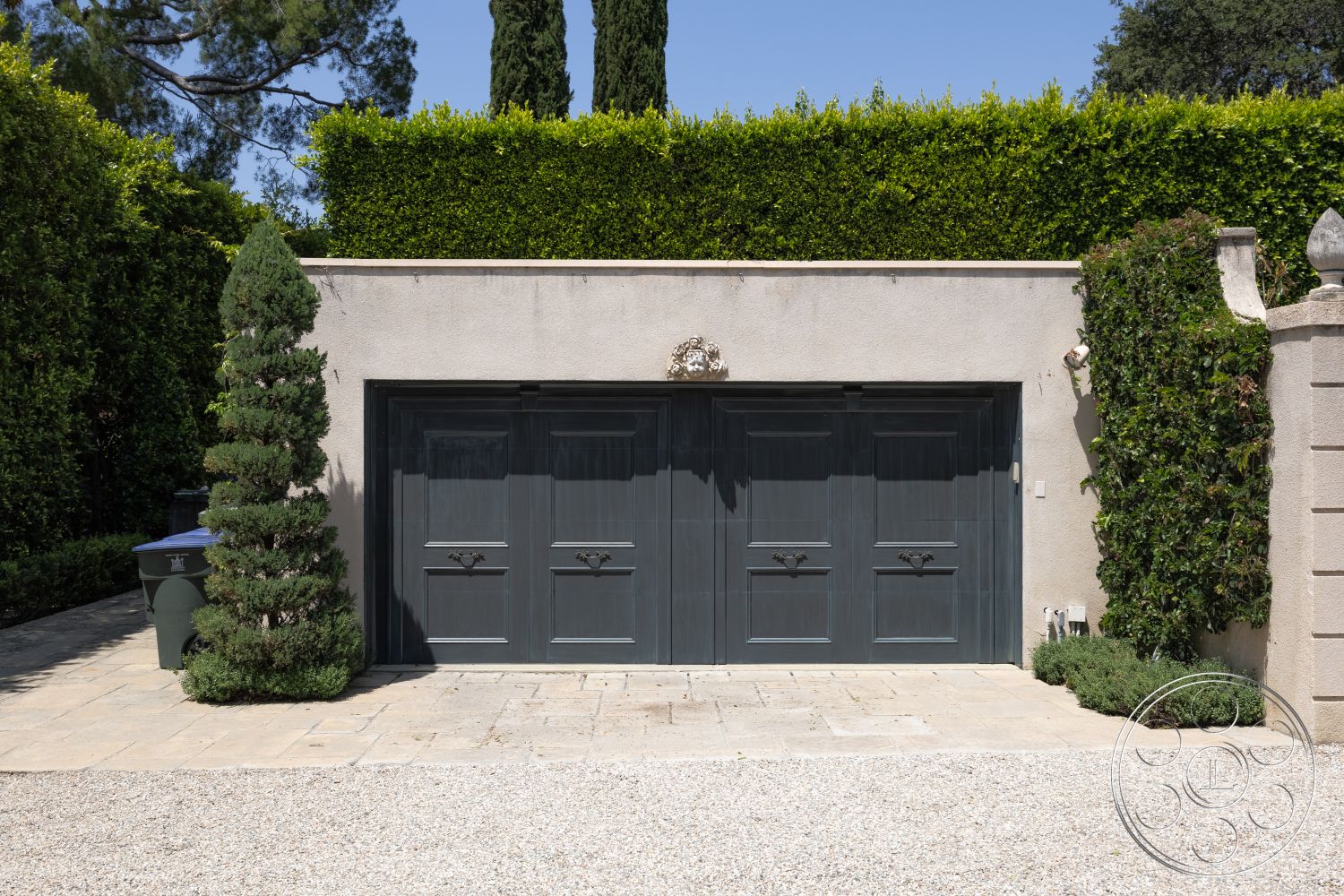 The Regent - gravel surface, landscaped hedges, flagstone paving, stucco wall finish, rounded topiary shrubs, exterior garage facade, flat concrete driveway, gray wooden garage doors, greenery border, decorative stone wall accents