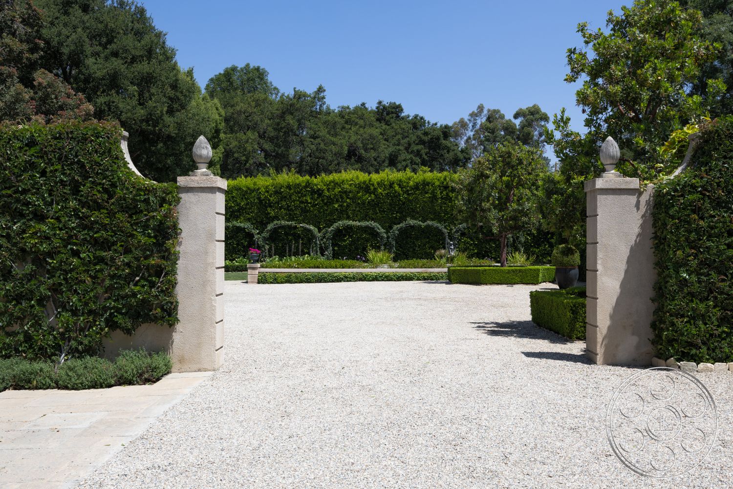The Regent - exterior, gravel driveway, formal garden layout, surrounded by trees, lush green foliage, stone entrance pillars, exposed aggregate concrete path, hedge perimeter landscaping, manicured garden beds, ornamental potted plants, smooth stucco wall, cobblestone patio area, curved garden pathways, architectural gate design, urbane setting, sun-drenched landscape