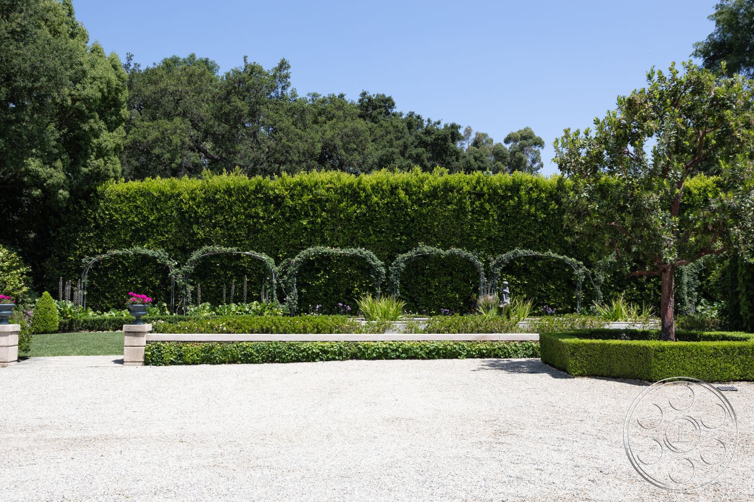 The Regent - fenced garden area, gravel driveway, ornamental trees, outdoor relaxation space, hedge privacy wall, natural greenery backdrop, symmetrical garden layout, stone garden bench, arched trellis feature, flowering plants garden bed, manicured lawn area, exterior landscape setting, potted plants accents, decorative stone borders, stone pathway elements