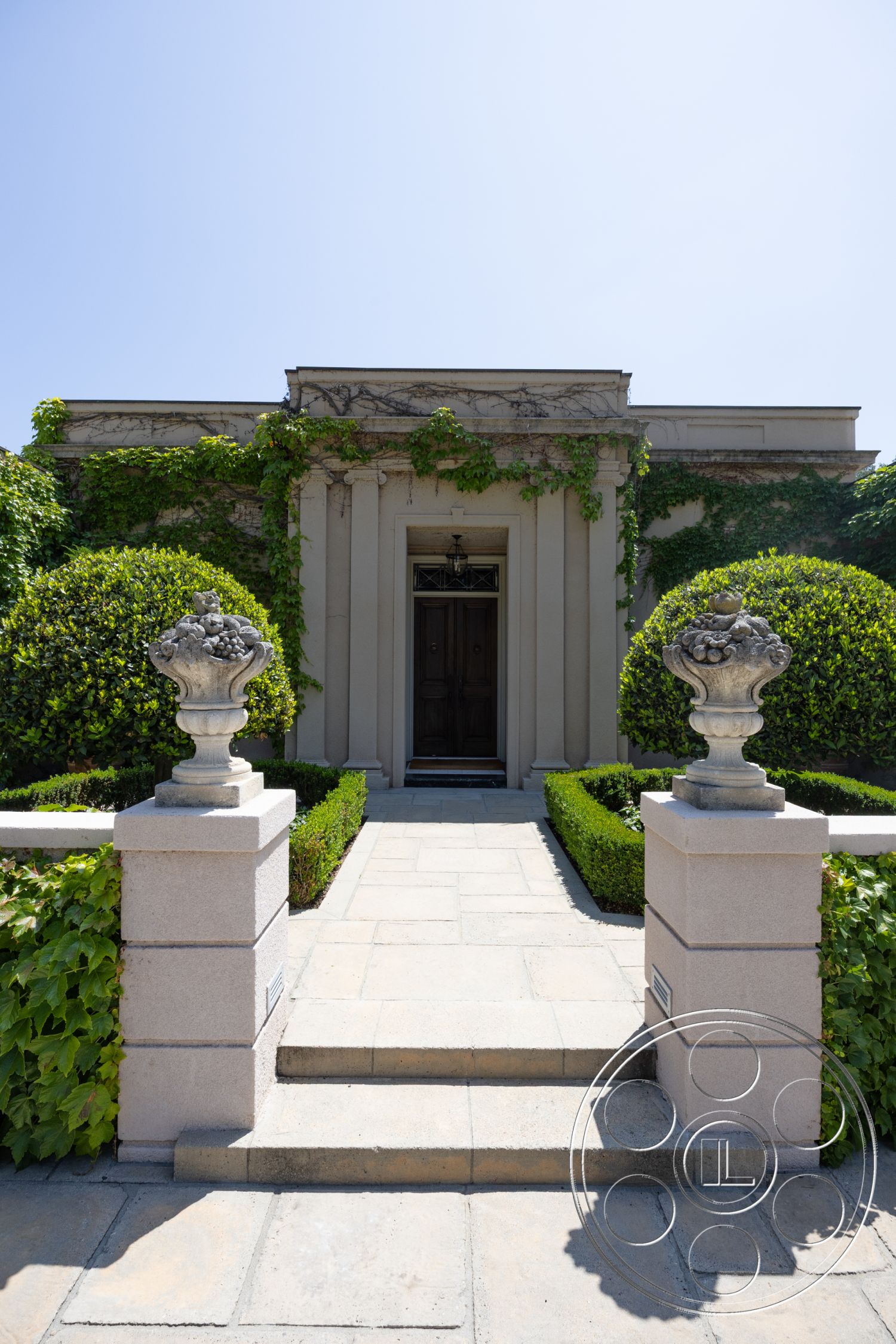 The Regent - formal garden setting, lush green shrubbery, exterior landscaped garden, exterior stone pathway, rectangular stone pavers, exterior ivy-covered walls, decorative stone wall accents, exterior stone steps, exterior concrete base, exterior brick planters, exterior manicured hedges, exterior double wooden doors, classical stone urn planters, exterior columned entryway, symmetrical façade, exterior light fixture