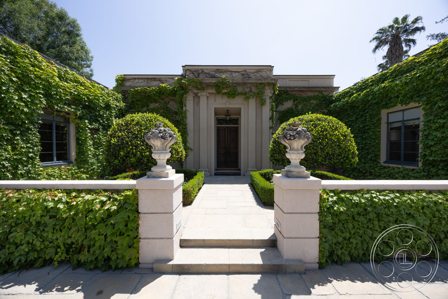 The Regent - exterior, modern entryway, classic architectural style, decorative stone railing, framed glass windows, stone walkway with concrete borders, landscaped garden with ivy-covered walls, ornamental stone urns, rectangular hedges, concrete steps leading to door, tropical palm tree landscaping, traditional columned portico, symmetric front elevation, lush greenery surrounding home, detailed molding on entry, geometric shapes in landscaping