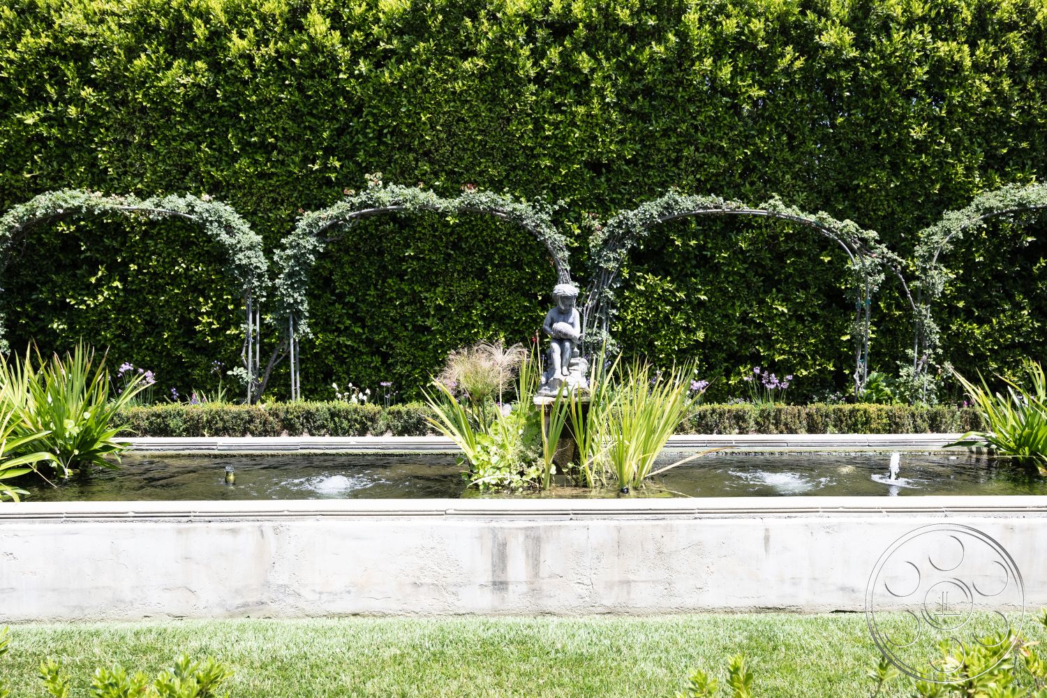 The Regent - lush greenery backdrop, exterior garden setting, concrete pond border, rectangular water feature, textured stone fountain, landscape with ornamental grasses, metal arch trellis with vines, polished stone edging, surrounding hedges and flower beds, architectural statue focal point