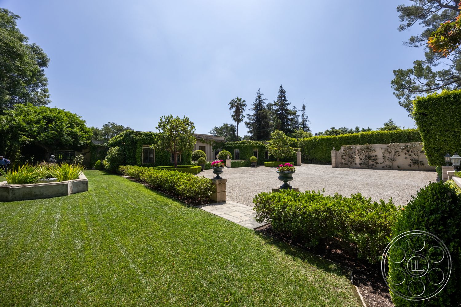 The Regent - outdoor living space, lush green lawn, tall hedges, gravel driveway, landscaped flower beds, stone planter boxes, exterior garden setting, decorative urns in garden, paved patio area, mature trees in background, hedge-lined pathways, modern villa style