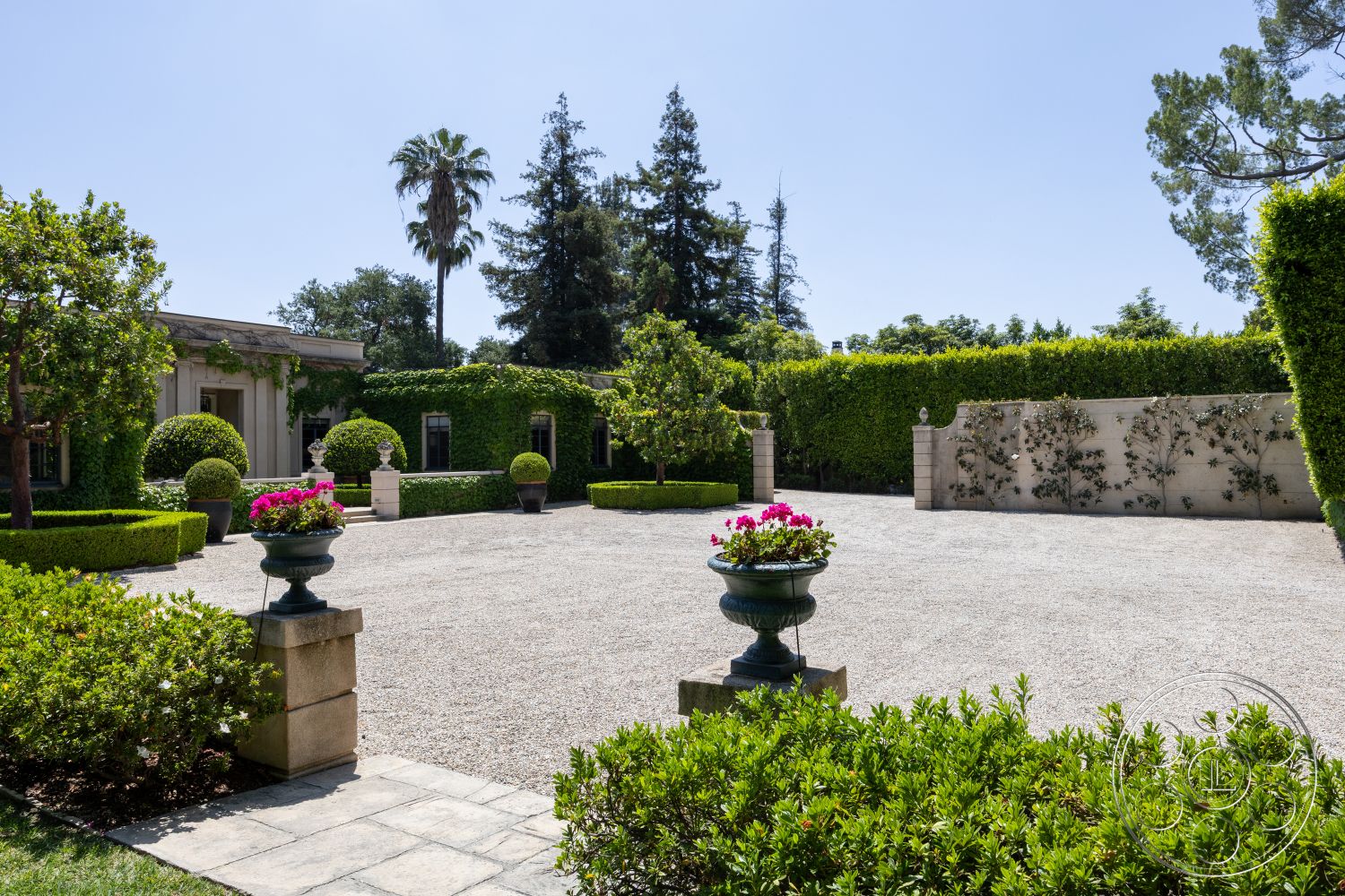 The Regent - stone wall, concrete planters, patio seating area, sunny courtyard, gravel driveway, tropical palm trees, exterior landscaped garden, suburban front yard, lush greenery surrounding, hedged garden border, circular topiary shrubs, granite coping stones, flowering plants in pots, archway landscape design, outdoor decorative elements, elegant outdoor aesthetic, Mediterranean-style home exterior, ornamental garden, flower beds with annuals, exposed light fixture, concrete terrace flooring