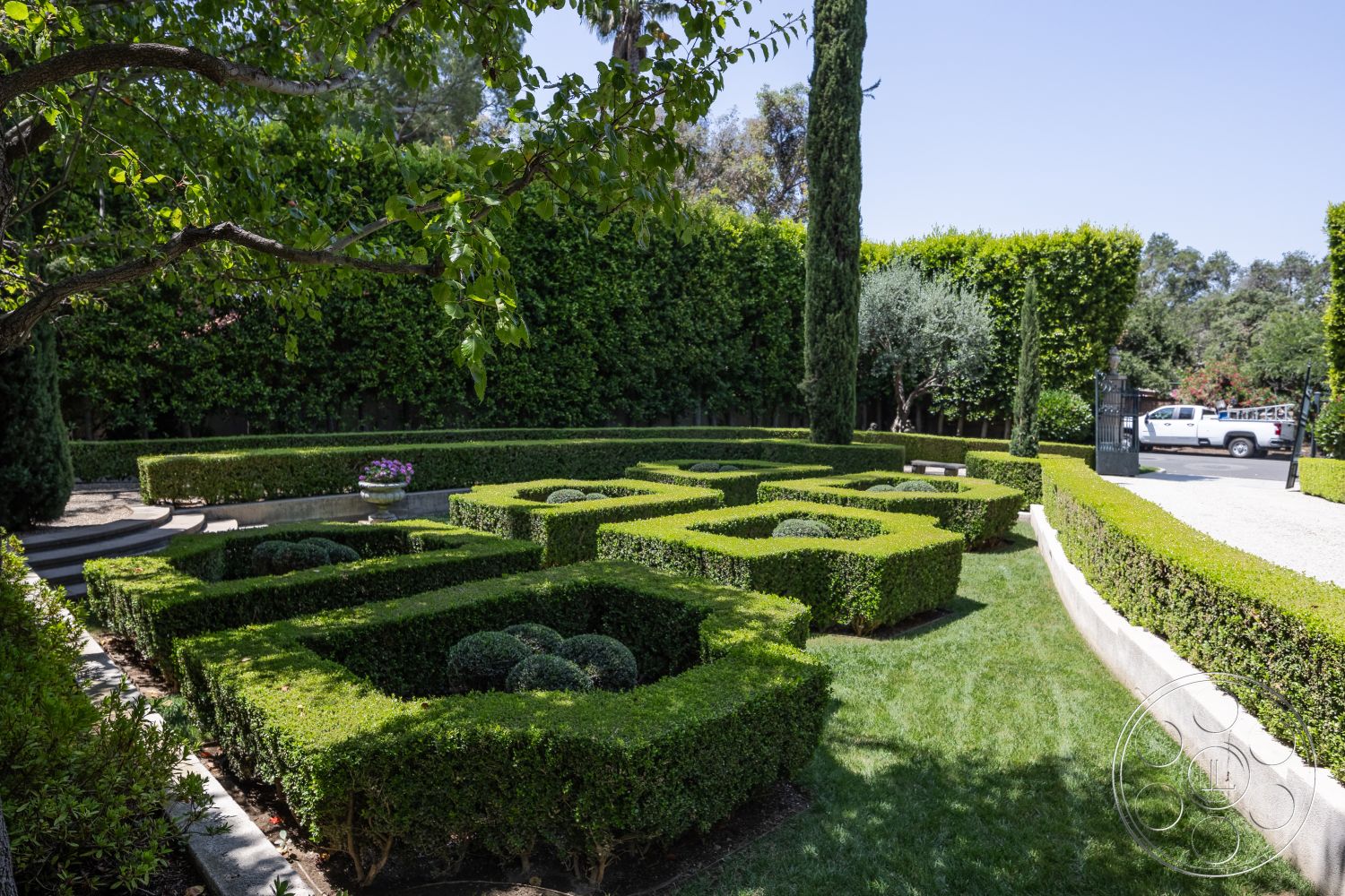 The Regent - exterior landscaping, tall cypress trees, gravel driveway, ornamental trees, manicured hedges, formal garden layout, circular stone planters, landscape design elements, urban garden setting, boxwood shrubs, curved stone pathway, foreground green lawn, sunlit garden space, vibrant flower accents, smooth stone edging, hedge maze pattern