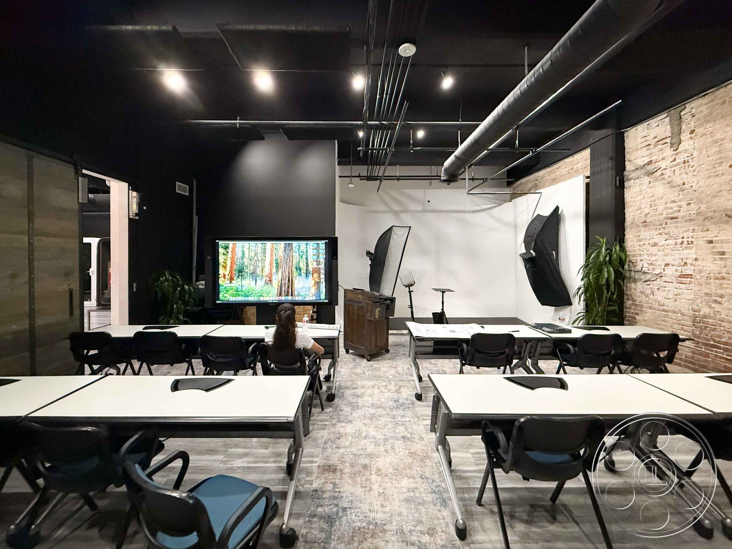 Office 124 - natural light interior, floor-to-ceiling windows interior, urban setting interior, exposed brick walls interior, black painted ceiling interior, modern classroom setup interior, whiteboard wall interior, folding tables classroom interior, black plastic stacking chairs interior, vinyl flooring with wood look interior, green plant decor interior, industrial-style lighting fixtures interior, creative workspace environment interior