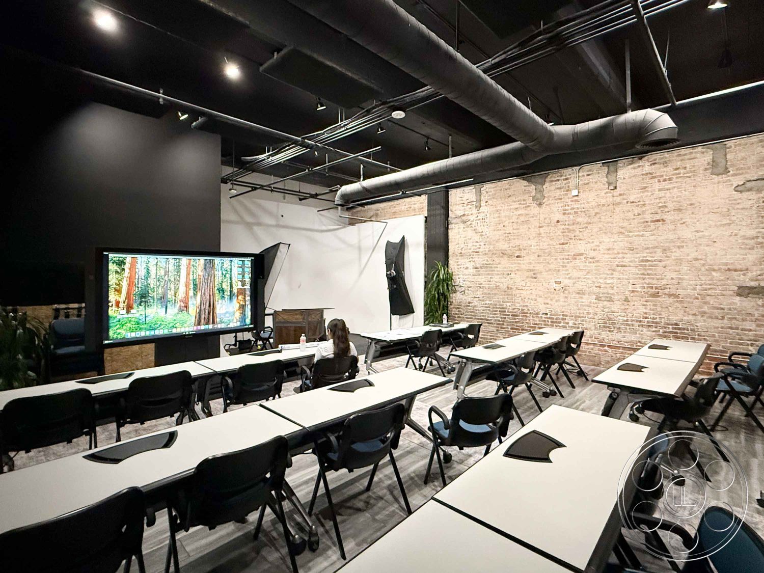 Office 124 - open floor plan, green indoor plants, large windows with natural light, urban office setting, interior brick accent wall, exposed brick wall treatment, black suspended ceiling panels, industrial steel ductwork, modern conference room setup, rectangular conference tables, hardwood laminate flooring, stackable black chairs, ceiling-mounted projection screen, large wall-mounted display monitor, minimalist decoration style, neutral color palette with black accents, dimmed lighting fixtures, tech-enabled meeting space, collaborative workspace design