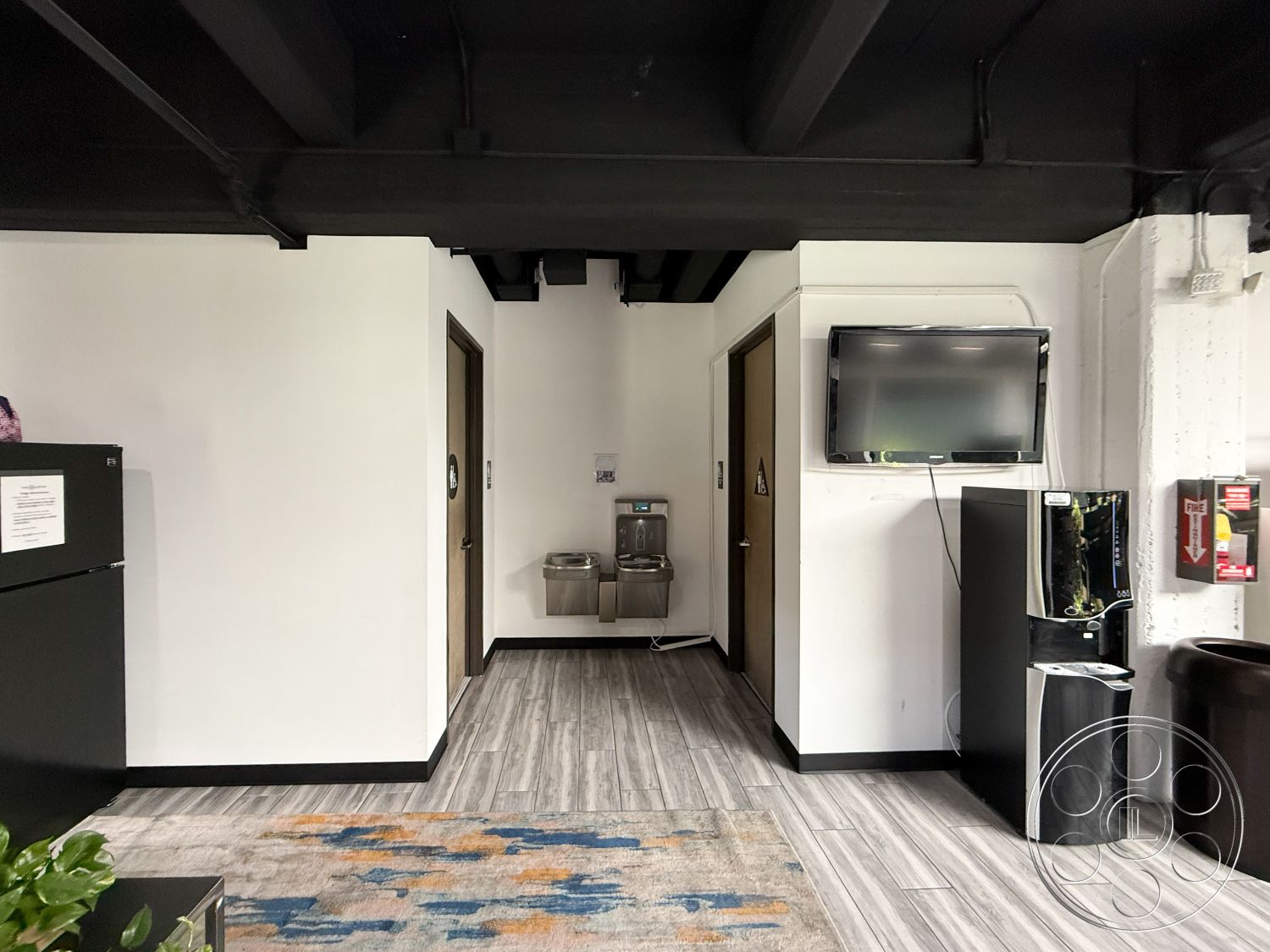 Office 124 - interior flooring - textured wood-look vinyl planking, interior wall treatment - white painted walls, exposed ceiling - black painted wooden beams, kitchen appliances - black refrigerator, reception area features - flat-screen wall-mounted television, water fountain - stainless steel drinking fountain, door material - wooden doors with dark finish, rug - area rug with abstract blue and orange patterns, interior furnishings - black plastic waste bin, interior ambiance - modern office design