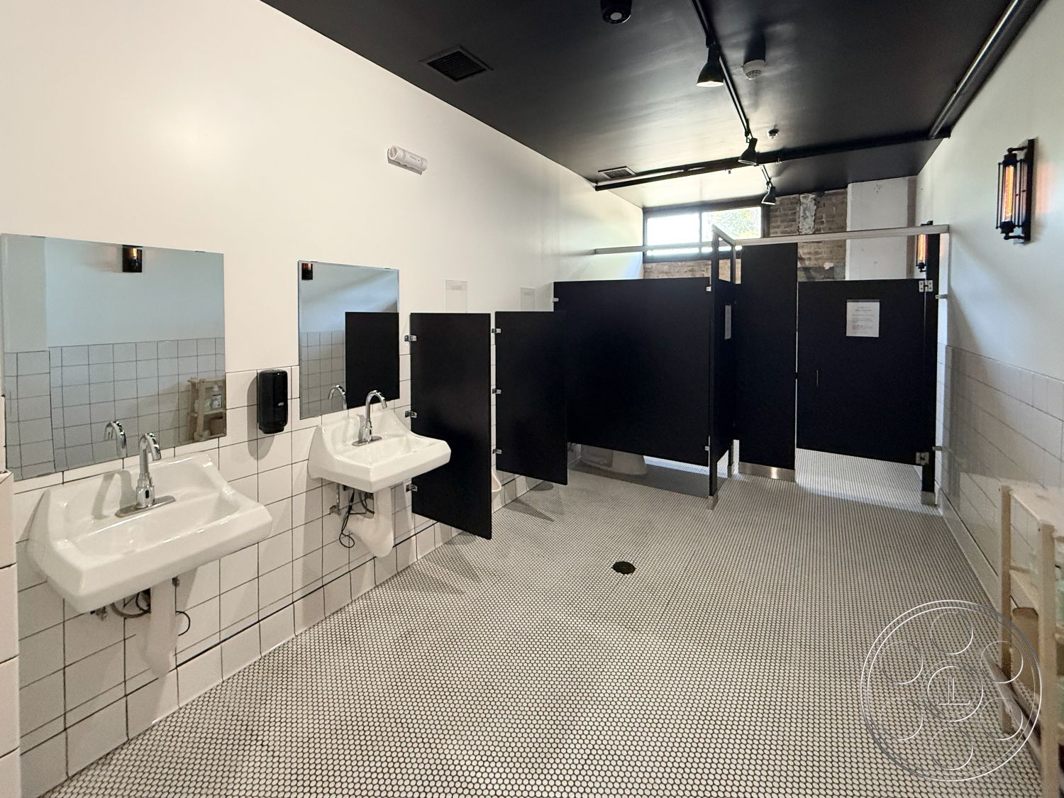 Office 124 - clean lines, minimalist design, functional layout, modern fixtures, bright natural lighting, black ceiling, interior bathroom, white painted walls, wall-mounted faucets, industrial-style lighting, white ceramic sinks, black privacy stalls, white subway tile wainscoting, black hexagonal tile flooring, exposed brick accents